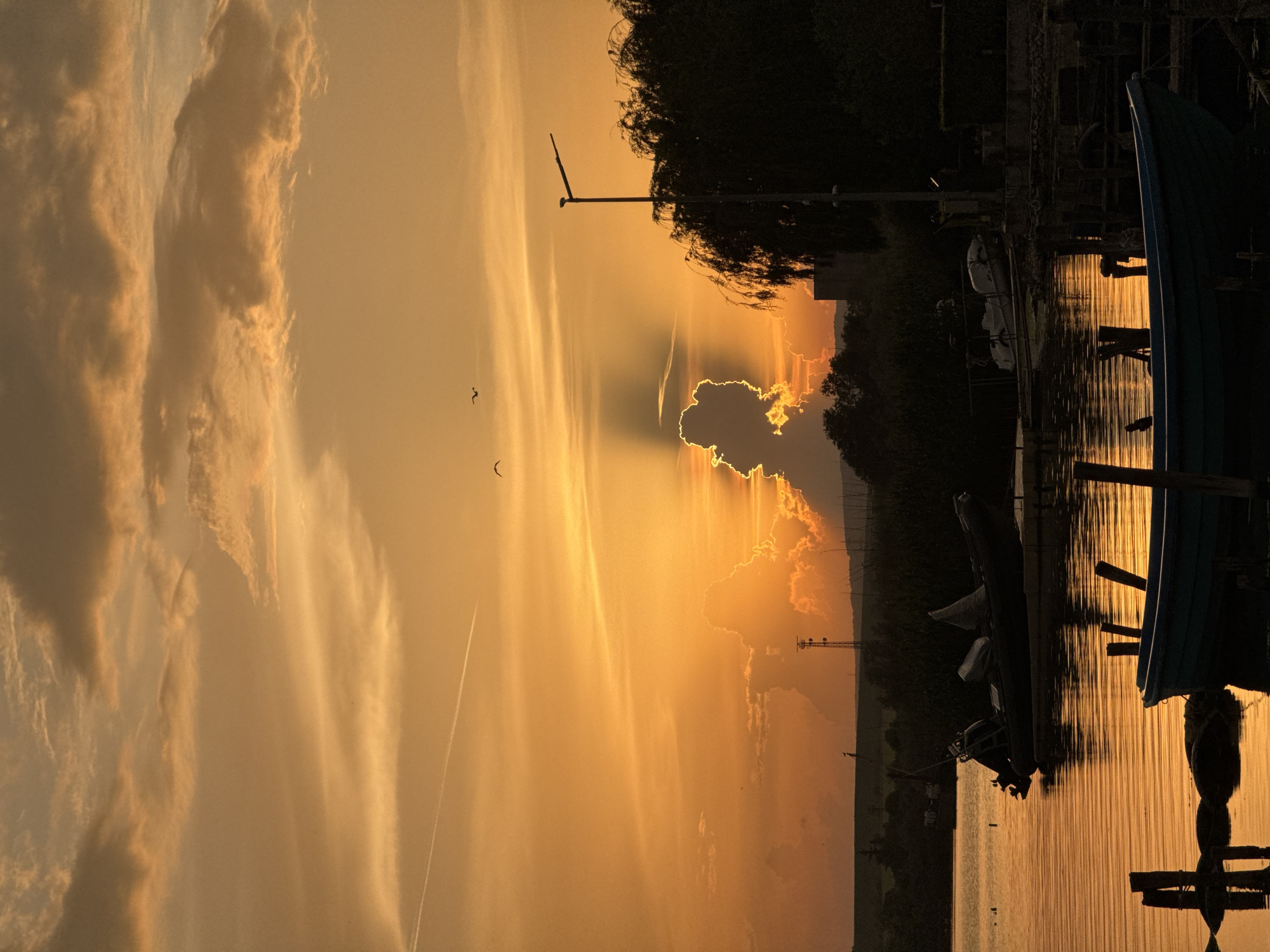 The image captures a sunset over a body of water, likely near Казашко, Bulgaria. A pier extends into the water, with several boats docked along it. One boat, partially visible on the right, is a darker color. The sunset casts an orange hue over the scene, with reflections visible on the water's surface. Silhouetted trees and structures are visible along the shoreline. There appear to be two birds flying in the sky.