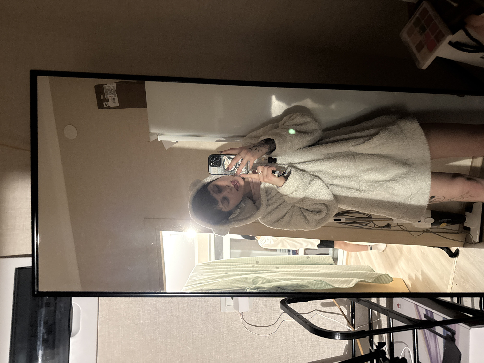 An individual is captured in a mirror selfie within an indoor residential setting in Saint Petersburg, Russia. The primary subject, a person with short dark hair, light complexion, and visible tattoos on the left arm and thigh, is wearing an off-white, fluffy hooded garment featuring bear ears. They hold a black smartphone in their right hand, angled to take the photo, while their left index finger rests near their mouth and their tongue is slightly extended. Rings are visible on the fingers, and a black key fob hangs from the phone.
The mirror's reflection reveals details of the surrounding room. To the primary subject's left, a brightly lit adjacent room or doorway is visible, showing a second individual partially standing on light wooden flooring, dressed in a light-colored top, dark shorts, and white socks. A white stick vacuum cleaner with its cord wrapped is positioned upright near this doorway. A green garment with a striped and dotted pattern hangs on a rack. To the primary subject's right, a white refrigerator stands beneath a light beige wall that features a circular white fixture. On top of the refrigerator, a brown cardboard box with visible QR codes and Cyrillic text, including "ПОЧТЫ РОССИИ" (Russian Post) and "A2", is present.
Outside the mirror, to the left, a light-colored wall with a power outlet, a white charging adapter, and a black cable is visible, alongside part of a black metal rack. In the lower right corner, a multi-compartment makeup palette rests on a surface next to a white paper bag. The scene is illuminated by artificial indoor lighting.