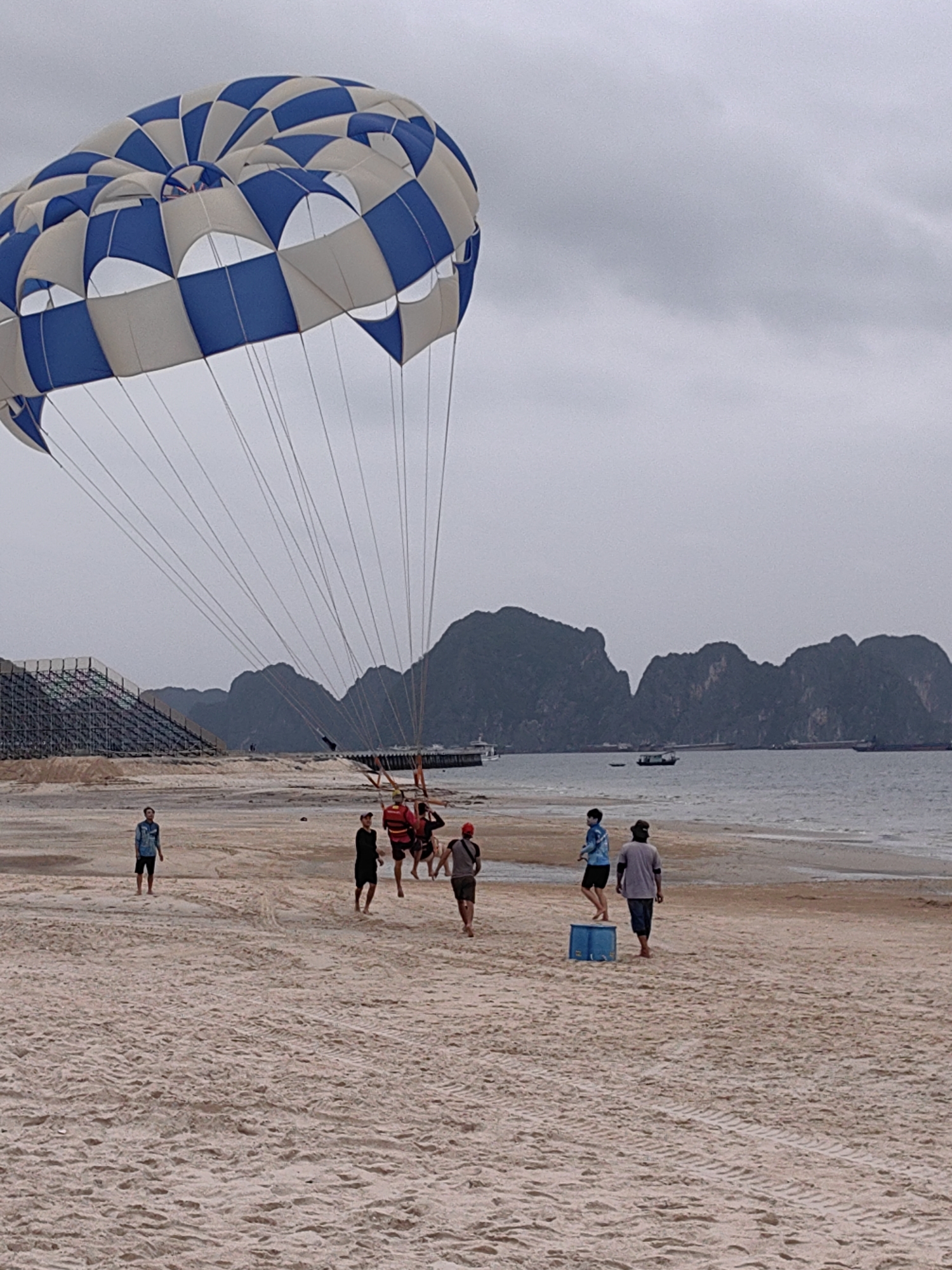 This image captures a vibrant parasailing scene on a sandy beach in Bai Chay Ward, Vietnam. The main subject is a large parasail, prominently displayed in the upper left, featuring a distinctive blue and white checkered pattern.
Beneath the parasail, several individuals are gathered on the light-colored sand. A group of people are actively engaged with the parasail's rigging, with one person visible in a red life vest, suggesting preparations for launch or a recent landing. Other individuals are walking or observing nearby on the beach. To the far left, a person stands alone, looking towards the activity. A blue plastic container rests on the sand in the mid-ground.
The setting includes a wide expanse of sandy beach, textured with footprints and what appear to be tire tracks. The ocean washes onto the shore on the right, where several boats and larger ships can be seen in the distance. The backdrop is dominated by dramatic, dark-colored limestone karsts, characteristic of coastal Vietnam, rising majestically from the water. On the left side of the image, a large, multi-tiered structure resembling bleachers or stadium seating is visible, adjacent to the beach.
The sky is uniformly overcast and grey, indicating a cloudy day. There is no visible text on any of the subjects or objects in the image. The overall impression is one of an adventurous recreational activity taking place on a scenic, yet somewhat muted, coastal day.