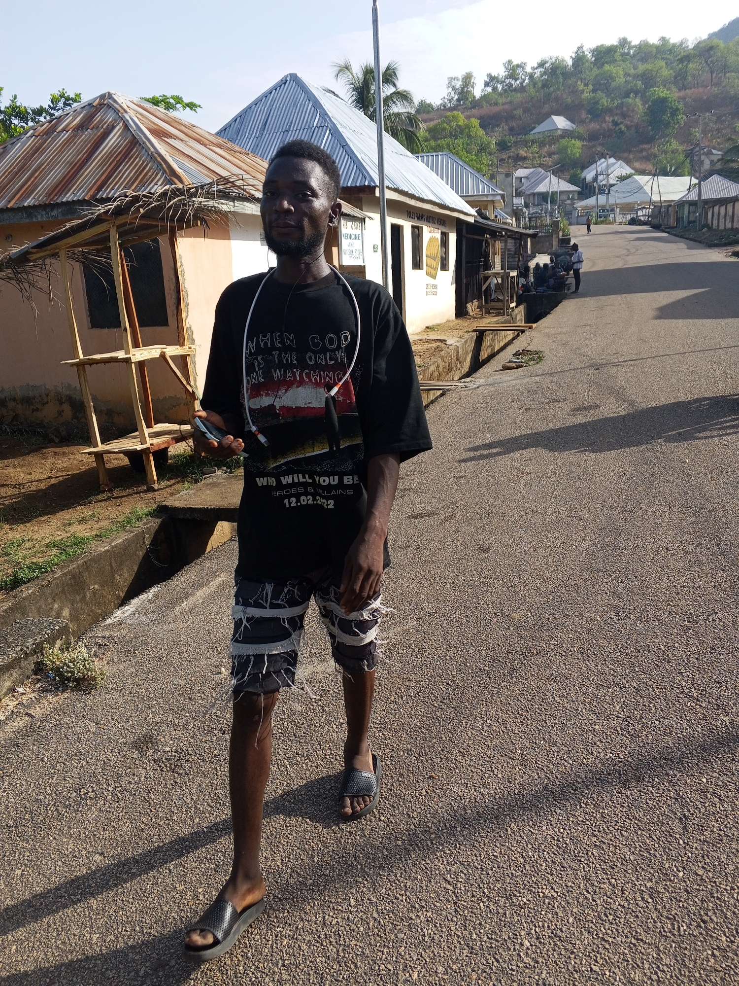 A young man walks down a paved road in Lokoja, Nigeria. He is wearing a black t-shirt with white text and an image on the front, fringed shorts, and sandals. He has earbuds around his neck and is holding a cell phone in his left hand. The road is lined with single-story buildings, some with corrugated metal roofs and open-air shops. There are a few people in the background. The scene is bathed in sunlight, with shadows cast across the road. A hillside with trees can be seen in the distance.