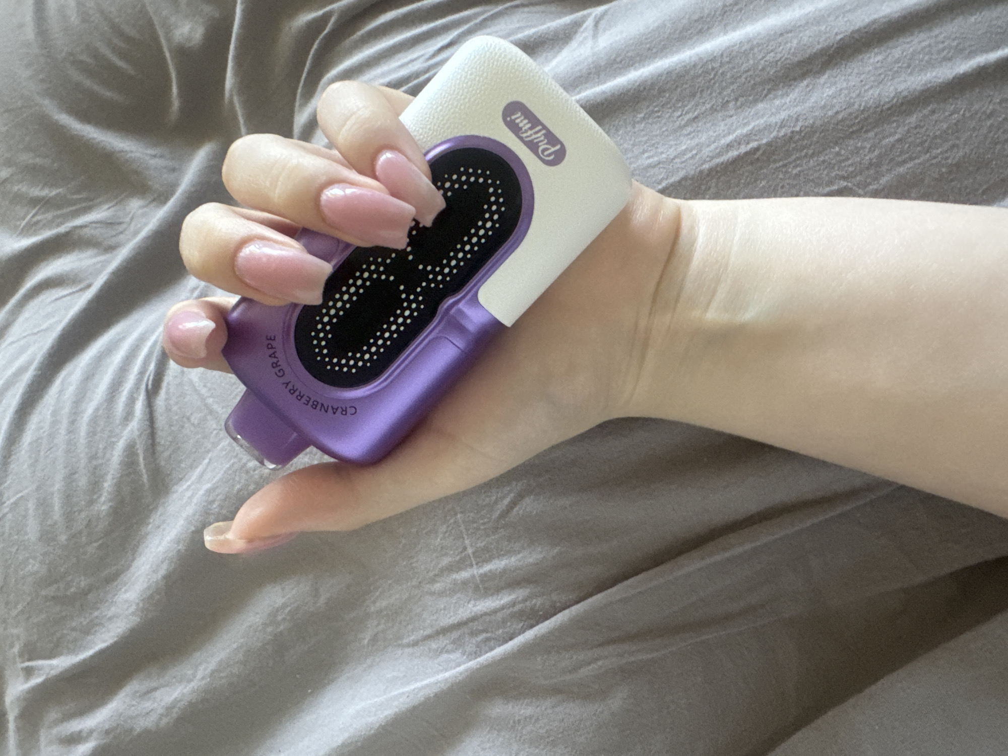 A person's hand with light skin and long, pink-colored artificial nails holds a purple and white electronic device. The device has a black mesh panel with white dots on the purple section and a logo that reads "Puffmi" on the white section. The device is positioned horizontally in the palm of the hand, with the fingers curled around it. The background is a gray, textured surface, possibly a bedsheet or fabric. The lighting is soft and diffused, casting subtle shadows. The image does not contain any information that allows for its location in Miass, Russia.