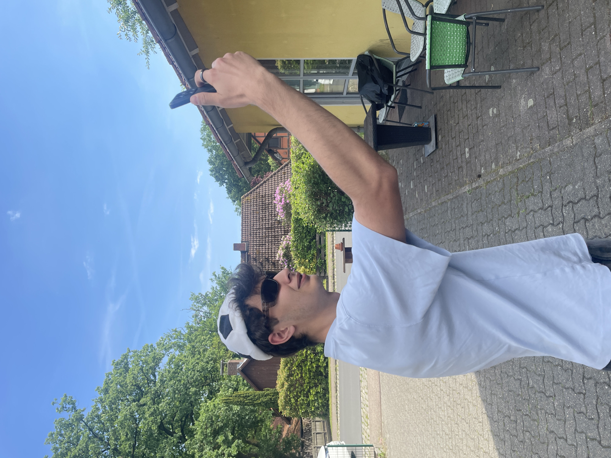 A young person with dark hair and sunglasses, wearing a white shirt and a white soccer-ball-patterned hat, stands on a paved surface outdoors, extending an arm upwards while holding a small black object in hand. The location appears to be in Wienhausen, Germany, judging by the surrounding architecture and vegetation. The background includes buildings, greenery, a brick-tiled roof, and a blue sky with scattered clouds. A yellow building with chairs and a black bag nearby stands behind the person.