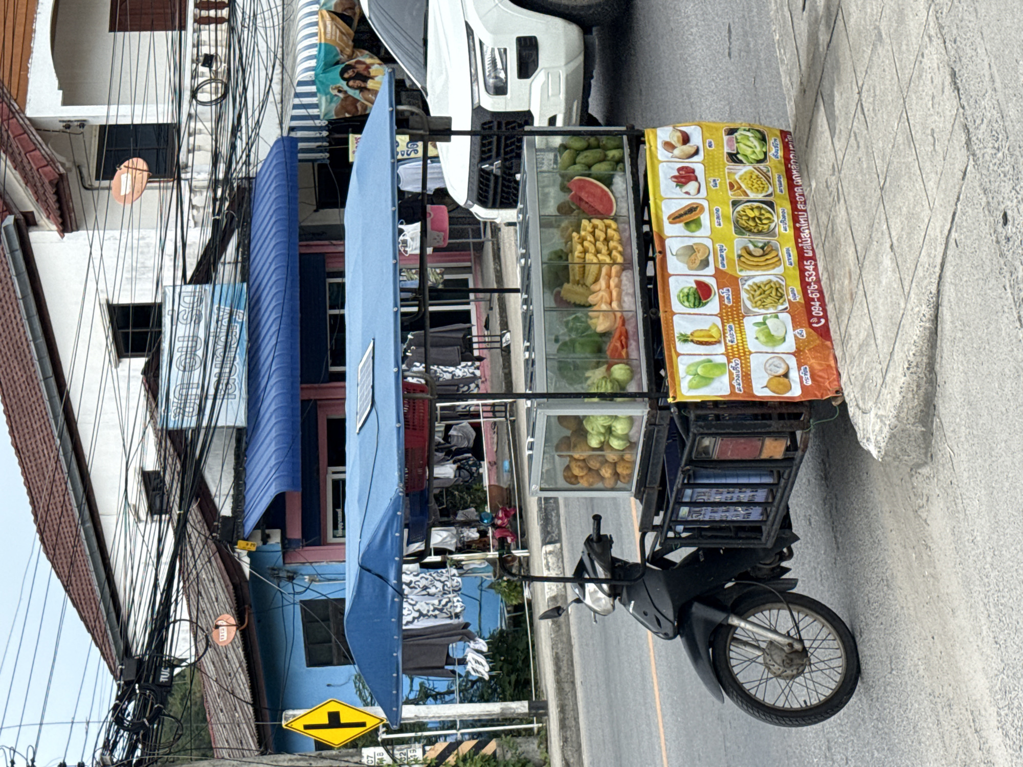 The image captures a street scene in Baan Bo Phut, Thailand, focusing on a fruit vendor's mobile cart. The cart is attached to a black scooter and is covered by a blue canopy. Inside a glass display, various fruits are arranged, including what appear to be guavas, pineapples, watermelons, and mangoes. A colorful banner displaying different fruits and a phone number is attached to the front of the cart. In the background, buildings with tiled roofs and electrical wires are visible. A white SUV is parked to the right of the fruit cart. The scene is set on a paved road with a concrete sidewalk.