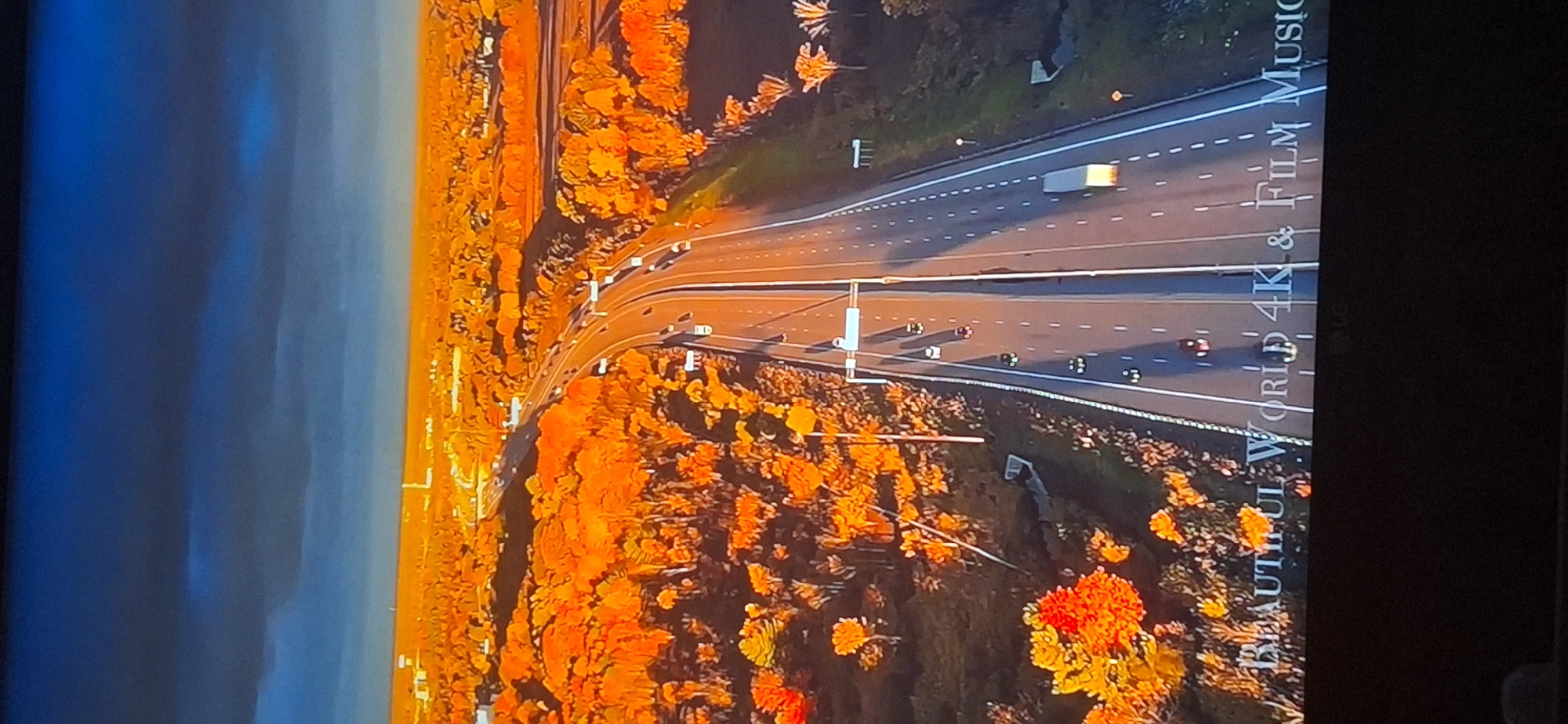 This is an aerial drone shot of a highway during sunset. The highway is flanked by trees with vibrant orange and yellow foliage, suggesting an autumn setting. Cars are visible on the highway, moving in both directions. The sky is a mix of dark blue and orange, with a sunset casting a warm glow over the landscape. In the distance, buildings and structures indicate an urban or suburban area. The text "BEAUTIFUL WORLD 4K & FILM MUSIC" is superimposed on the lower portion of the image. The location is not Bila Tserkva, Ukraine.