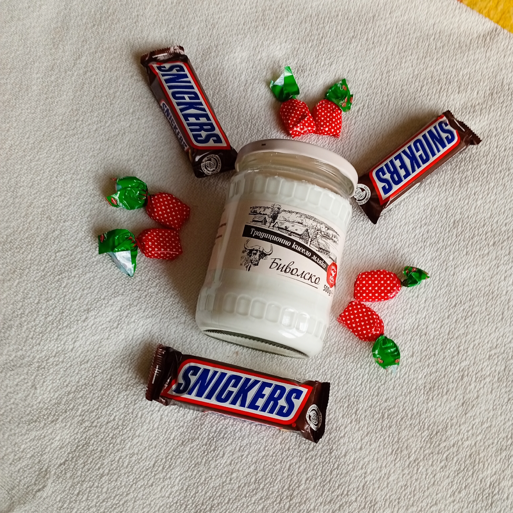 An overhead still-life image presents a collection of food items arranged on a textured, off-white fabric surface. At the center is a clear glass jar with a white plastic lid, containing an opaque white substance. The jar features a white paper label with a black and white illustration of a rural landscape, the Cyrillic text "Традиционно кисело мляко" (Traditional yogurt/sour milk), a buffalo head illustration, "Биволско" (Buffalo), and "500g," along with a small red circular seal.
Positioned radially around the central jar are three individual Snickers chocolate bars, each in its standard brown wrapper with blue and white branding. Interspersed with the Snickers bars are eight individual candies, each wrapped in red foil with white polka dots and twisted ends, with green foil at one end, resembling strawberries. The underlying surface is uniformly textured, consistent with a woven fabric like a towel or tablecloth. No discernible environmental features or landmarks are present to indicate a specific location within the city of Harmanli, Bulgaria. No people are visible. No actions or interactions are depicted.