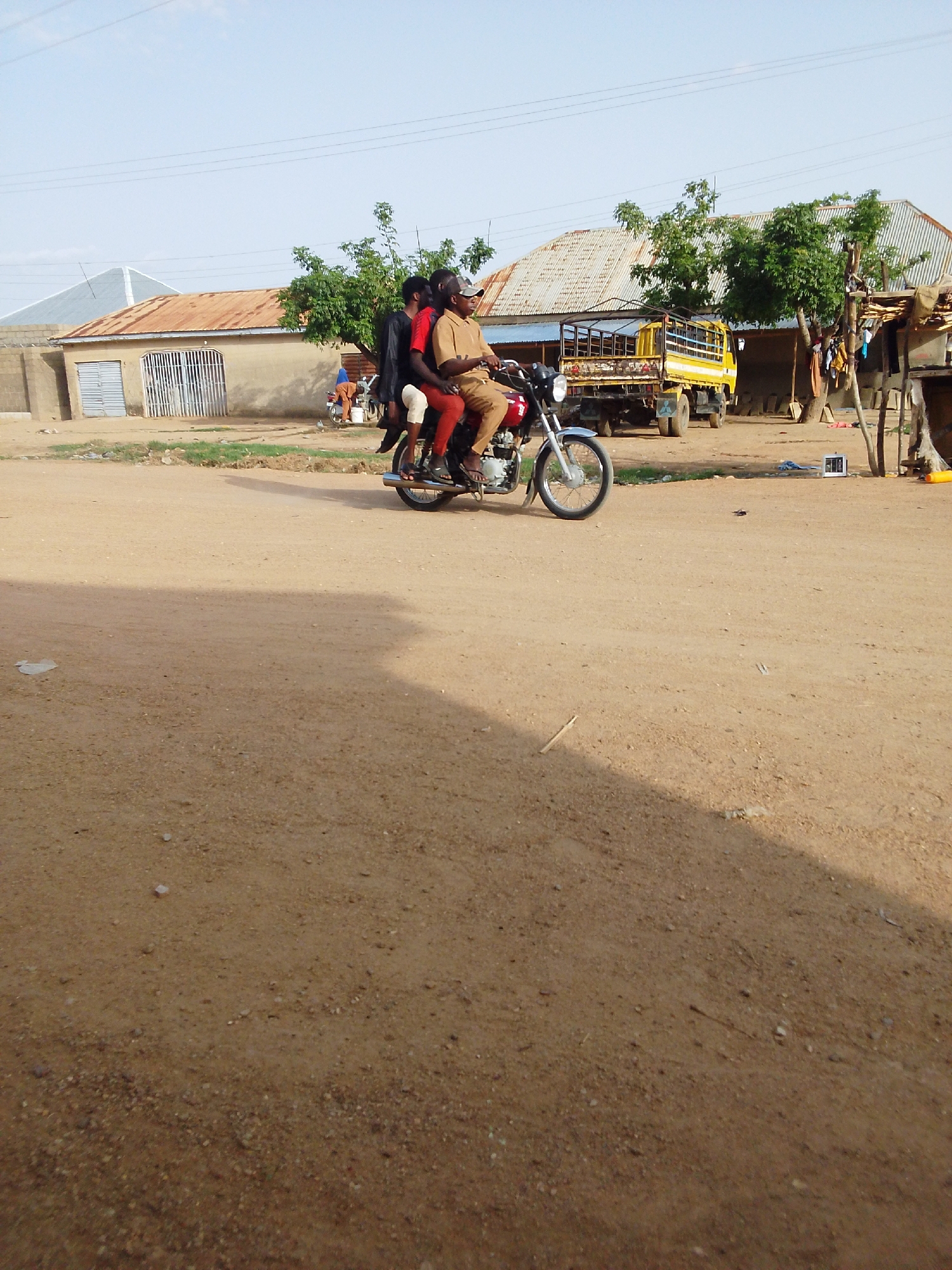 The image depicts a dirt road scene in Bauchi, Nigeria. Three individuals are on a red motorcycle driving on the road. The driver is wearing a brown shirt and cap, while the passenger is wearing a black jacket. There is a second passenger wearing a red shirt. In the background, there is a yellow truck, some buildings with corrugated metal roofs, and sparse trees. There are power lines in the sky. The ground is sandy and dusty.
