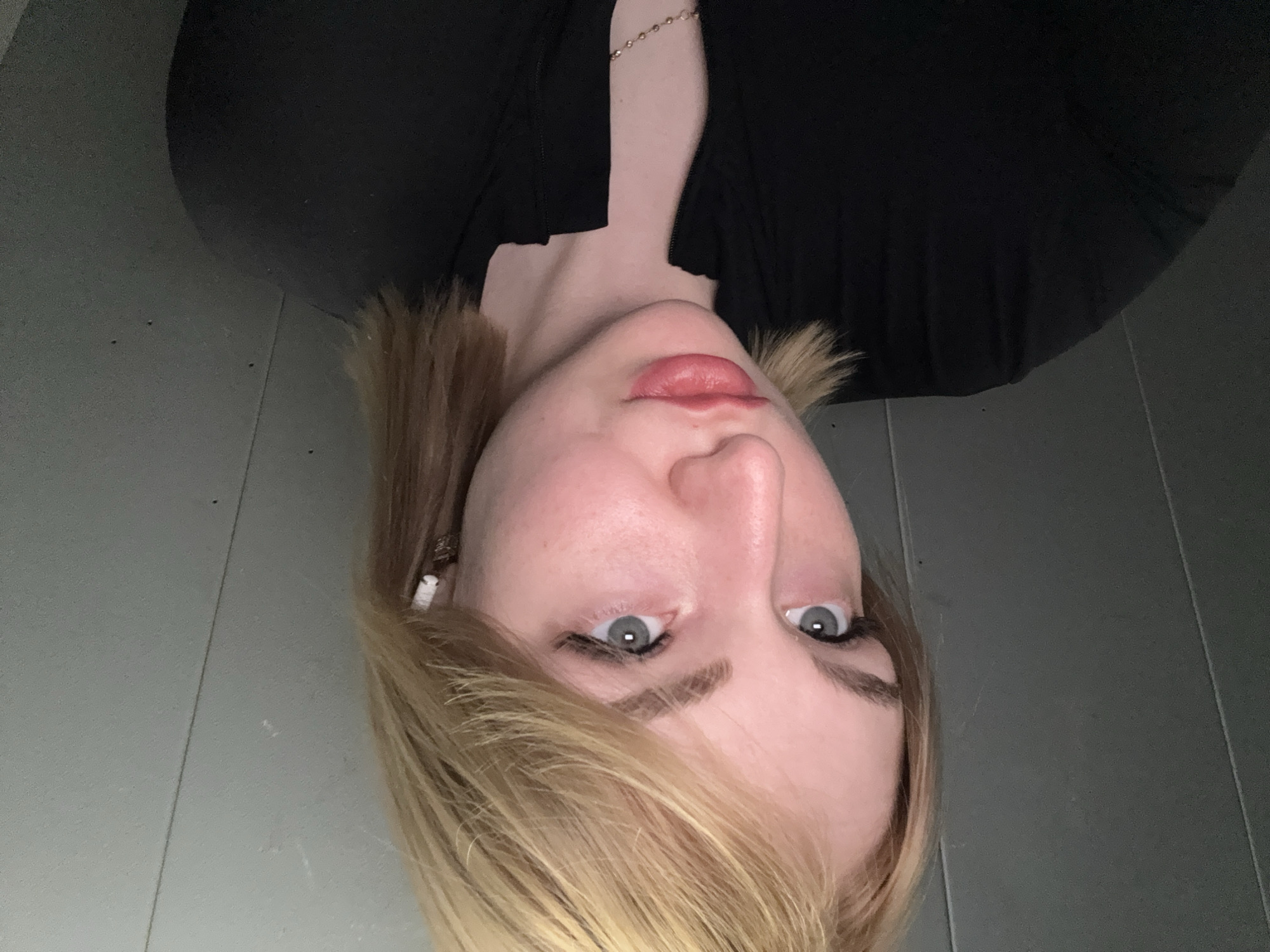 A young woman with blonde hair and blue eyes is pictured from the chest up, looking slightly to her left. She is wearing a black top and white earbuds. The background is a gray, textured wall. The location of this image is the city of Khimki, Russia.