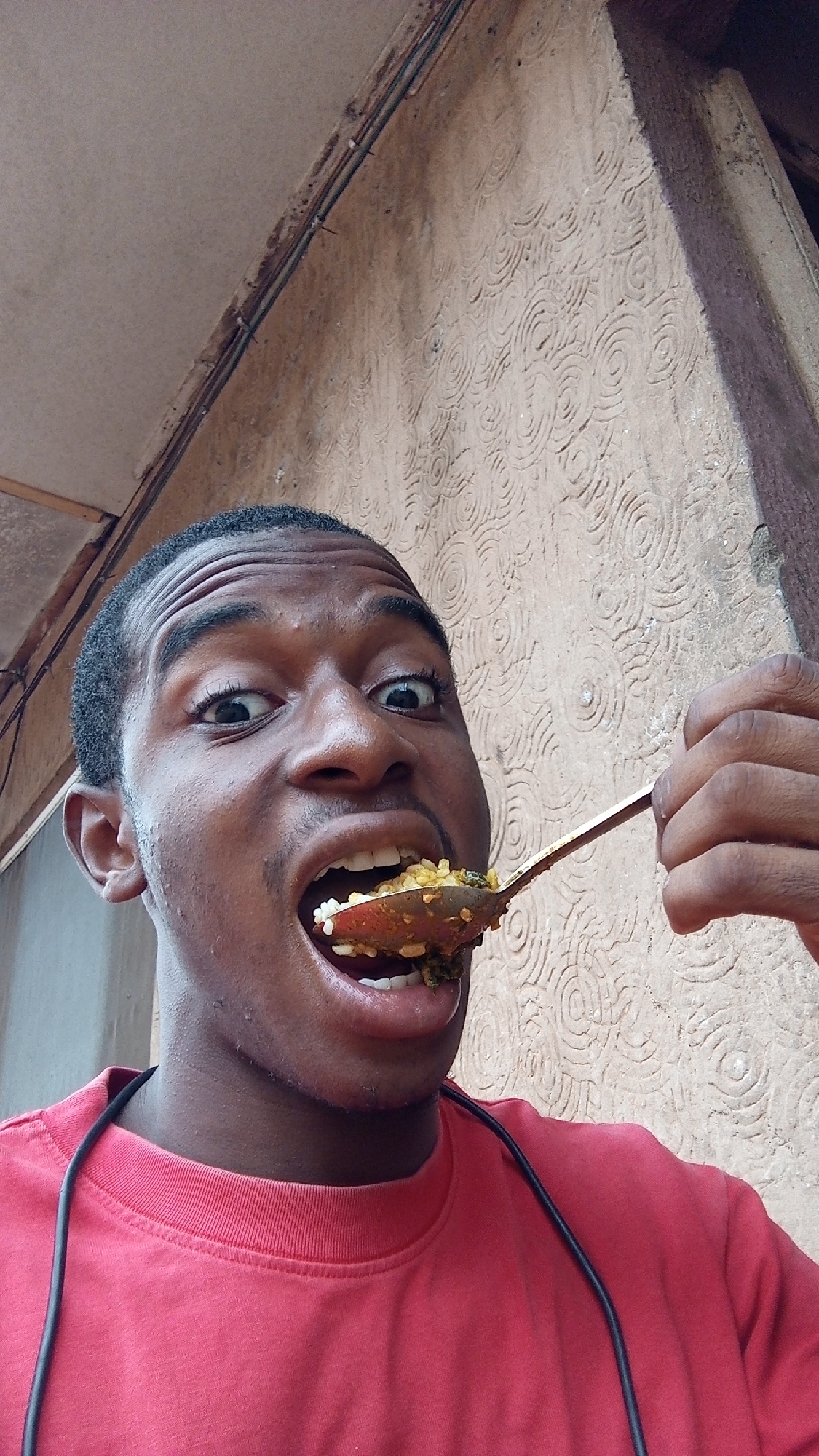 A close-up, low-angle shot captures a young Black male in Nkpor, Nigeria, in the act of eating. He looks directly at the camera with a wide-eyed, slightly exaggerated expression and an open mouth, conveying a playful or humorous mood. His forehead is wrinkled, contributing to the animated facial expression.
He wears a solid red long-sleeved shirt, and a black cord, possibly for headphones, is draped across his left shoulder and chest. In his right hand, he holds a gold-toned spoon, heavily laden with a mixture of food that appears to be rice combined with a dark green leafy stew or sauce. The spoon is positioned just at the entrance of his open mouth, suggesting he is either about to take a bite or has just done so.
The background features a light brown wall with a distinct, repeating pattern of embossed concentric circles, indicative of a decorative stucco or plaster finish. Above and to the left, a darker, smoother surface, possibly an architectural overhang or ceiling, is visible with exposed electrical wiring or cables running along its edge. On the right side of the frame, part of a rough concrete pillar or beam is present. The scene is brightly lit by natural light, indicating it is daytime. There is no visible text in the image.