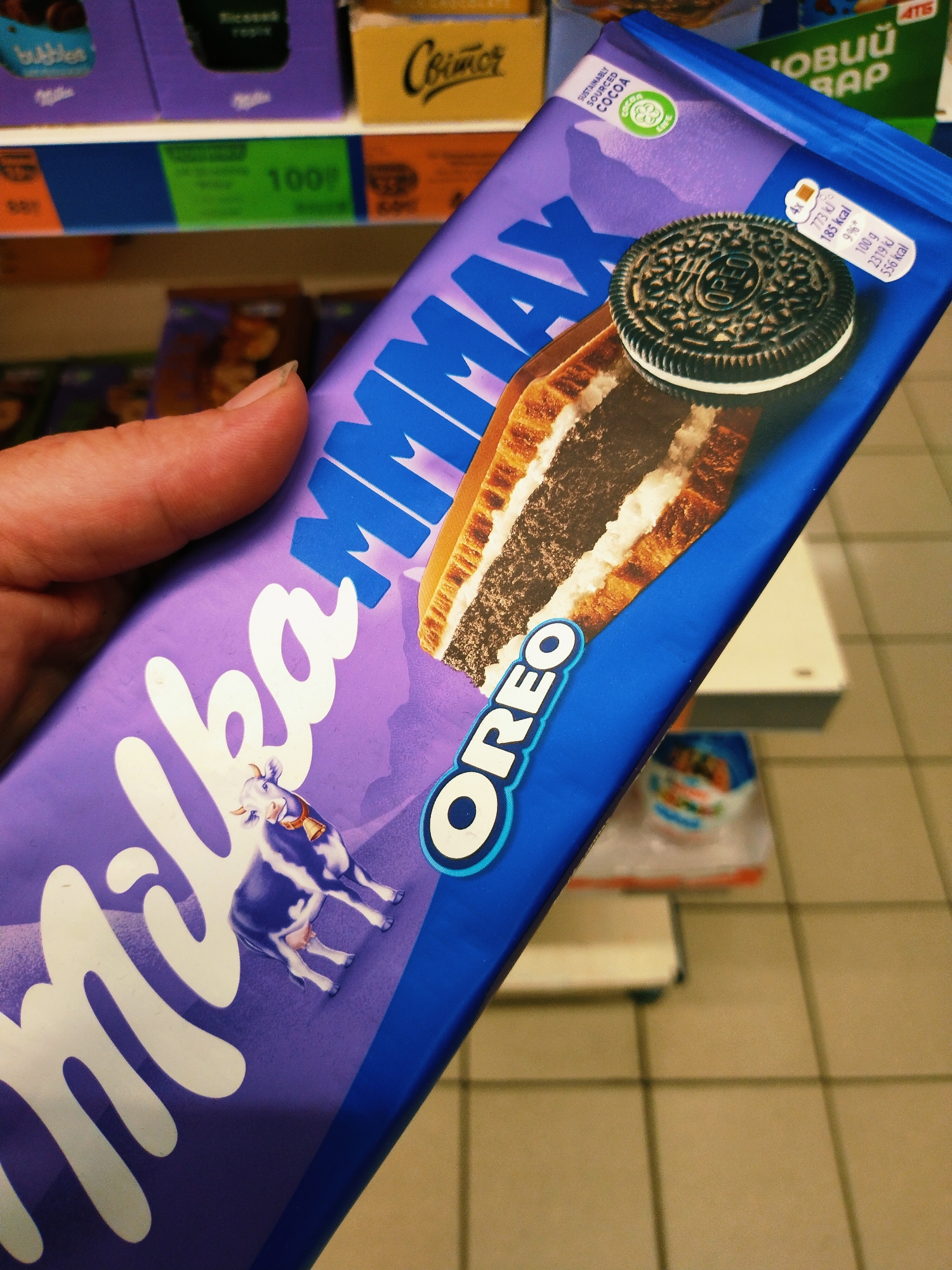 A hand holds a Milka Oreo Max chocolate bar in a store. The chocolate bar is purple with white lettering and features a large image of an Oreo cookie with a bite taken out of it, revealing the cream filling. The brand "Milka" is prominently displayed in large white script across the lower portion of the bar, with a stylized image of a Milka cow. The background shows shelves stocked with other confectionery items, suggesting the setting is a grocery store or supermarket. The floor is tiled with light brown squares. The image is taken from a slightly elevated angle. The scene is located in Horishni Plavni, Ukraine.