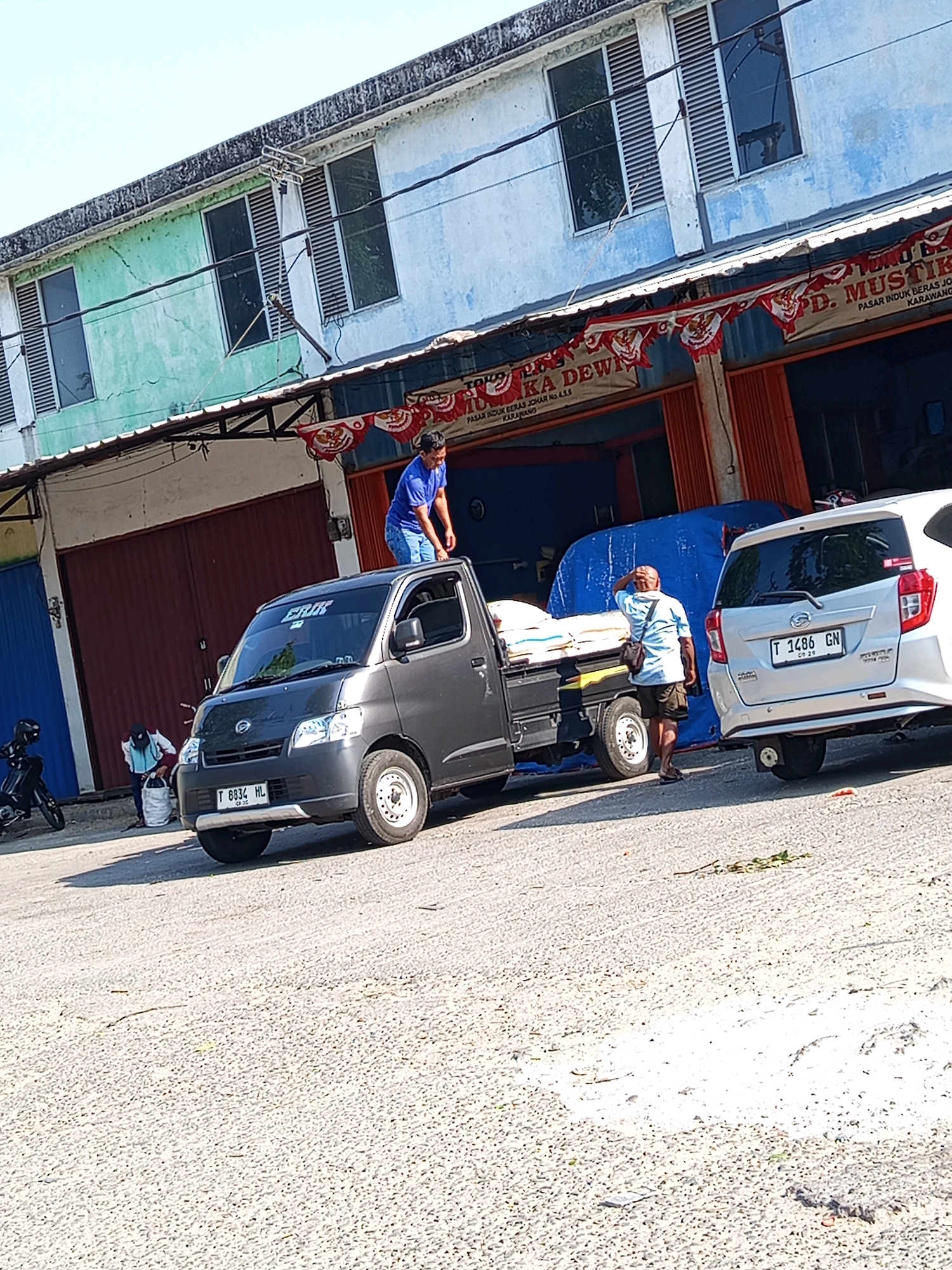 In Karawang, Indonesia, a gray pick-up truck is parked on a paved road in front of a two-story building with a weathered, blue and green facade. The building has multiple windows with louvers. A banner across the front of the building reads "TOKO BERAS MUSTIKA DEWI" with additional text below indicating it is a wholesale rice shop located in Pasar Induk Beras Johar, Karawang. Red and white pennants are strung across the front.
On the truck bed, several bags of rice are stacked. A man in a blue t-shirt stands on the roof of the pick-up truck, seemingly securing the load. Another man, wearing a light-colored shirt and dark shorts, stands near the truck, facing away from the camera with his hand on his head. To the right of the pick-up truck is a silver car. In the background, a motorcycle is parked to the left of the building. The scene is illuminated by bright sunlight, casting shadows on the ground.