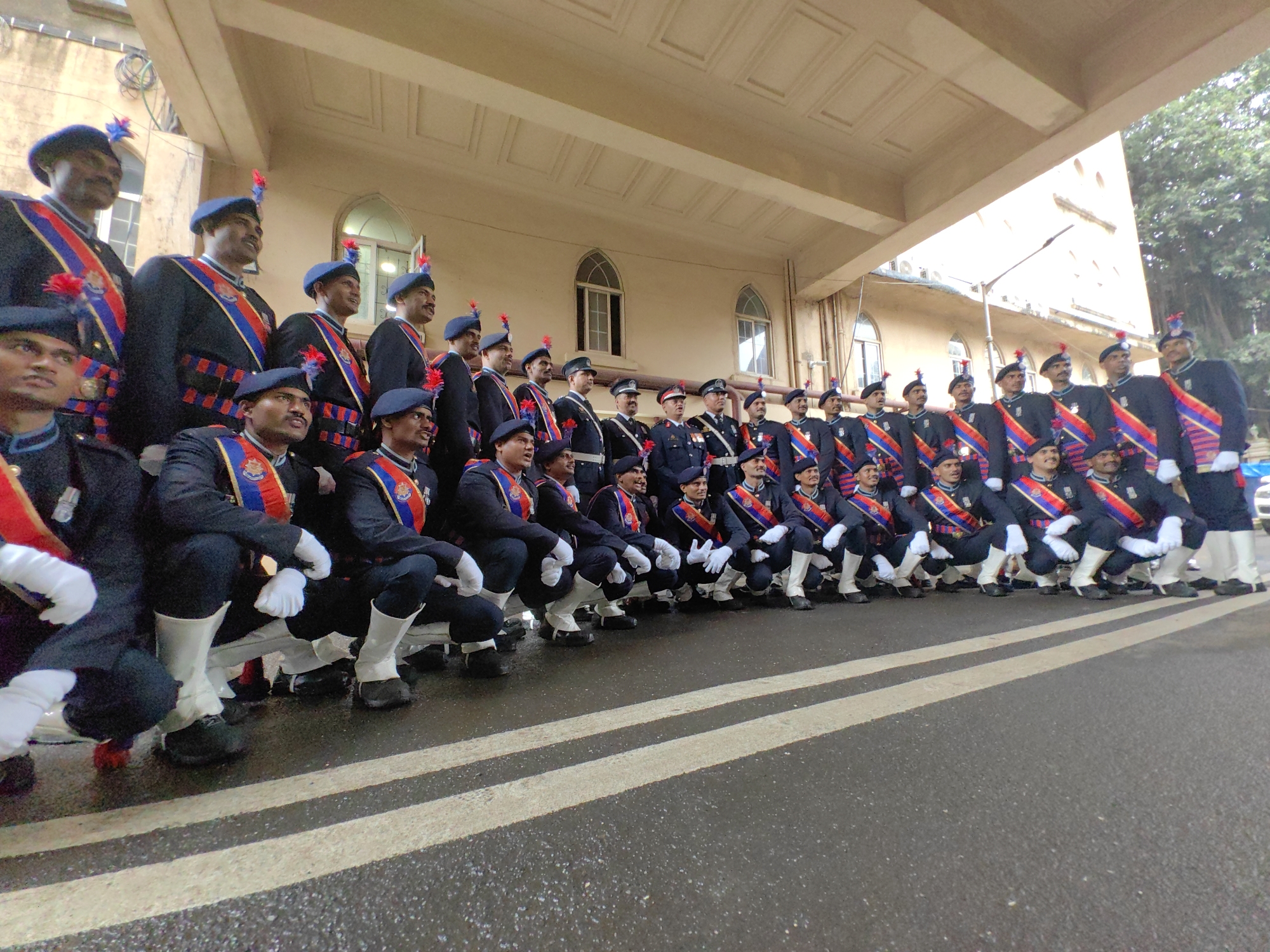 A group of men in uniform are posing for a photograph. They are all wearing dark blue jackets with red and blue sashes, blue berets, white gloves, and white gaiters over dark pants. Some are standing, while others are kneeling in a line, facing the camera. The men appear to be part of a ceremonial guard or military unit.
The background suggests a colonial-era architectural style, likely a public building with arched windows and a textured facade, possibly made of stone. The building's exterior is a warm, light brown or beige color, and it features a grand, covered entryway supported by columns. The sky above is overcast. The group is positioned on a wet, asphalt road marked with white double lines, indicating they are likely outdoors, possibly on a street or parade ground.
The overall scene evokes a formal and disciplined atmosphere. This image was taken in front of the **Victoria Terminus** (now Chhatrapati Shivaji Maharaj Terminus) in Mumbai, India, a UNESCO World Heritage Site known for its Victorian Gothic Revival architecture.