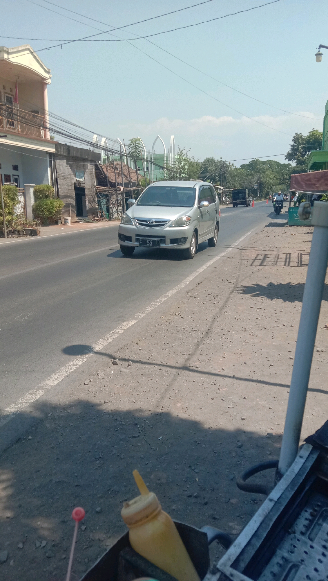 A silver minivan with license plate S 1607 TD is driving on a road in the city of Karang Anyar, Indonesia, on a sunny day. The minivan is on the right side of the road, traveling away from the camera. The road is paved and has a white line marking the lane. On the left side of the road, there is a building with a red flag on the balcony, and several trees. In the background, there are more buildings and trees, as well as a clear blue sky with a few clouds. The foreground shows a blurry image of a street cart with a yellow squeeze bottle and a red lollipop stick.