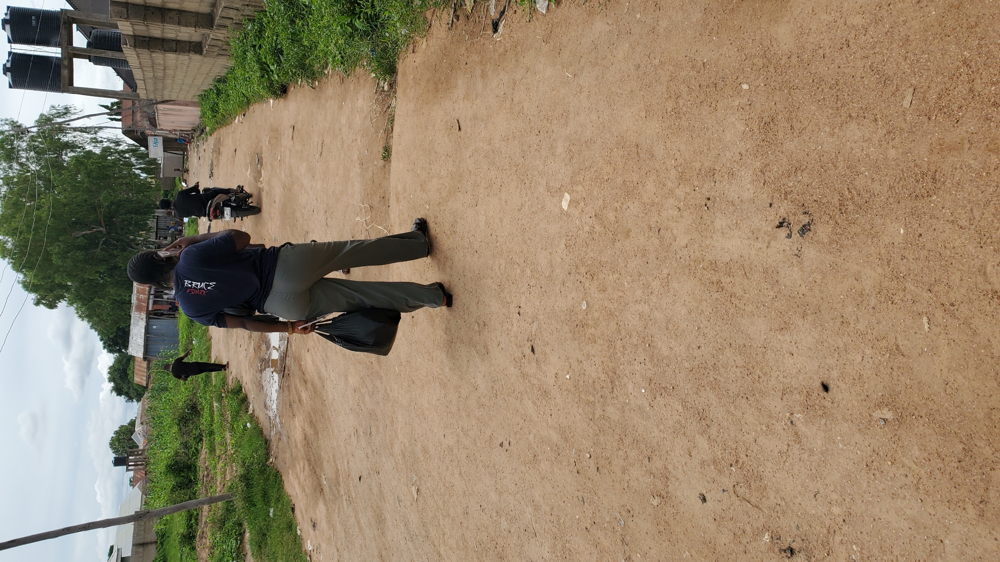 The image depicts a dirt road in Jalingo, Nigeria. In the foreground, a person is walking away from the camera, holding a black plastic bag in their left hand and speaking on a mobile phone. They are wearing a dark blue t-shirt with white lettering and olive green trousers. In the mid-ground, a person is riding a motorcycle, also moving away from the camera. Further in the background, another person is walking on the left side of the road, and beyond them, various buildings and vegetation are visible, including water tanks on a raised structure. The sky is overcast. The road is unpaved and appears to be in a residential or semi-developed area, with some puddles of water indicating recent rain.