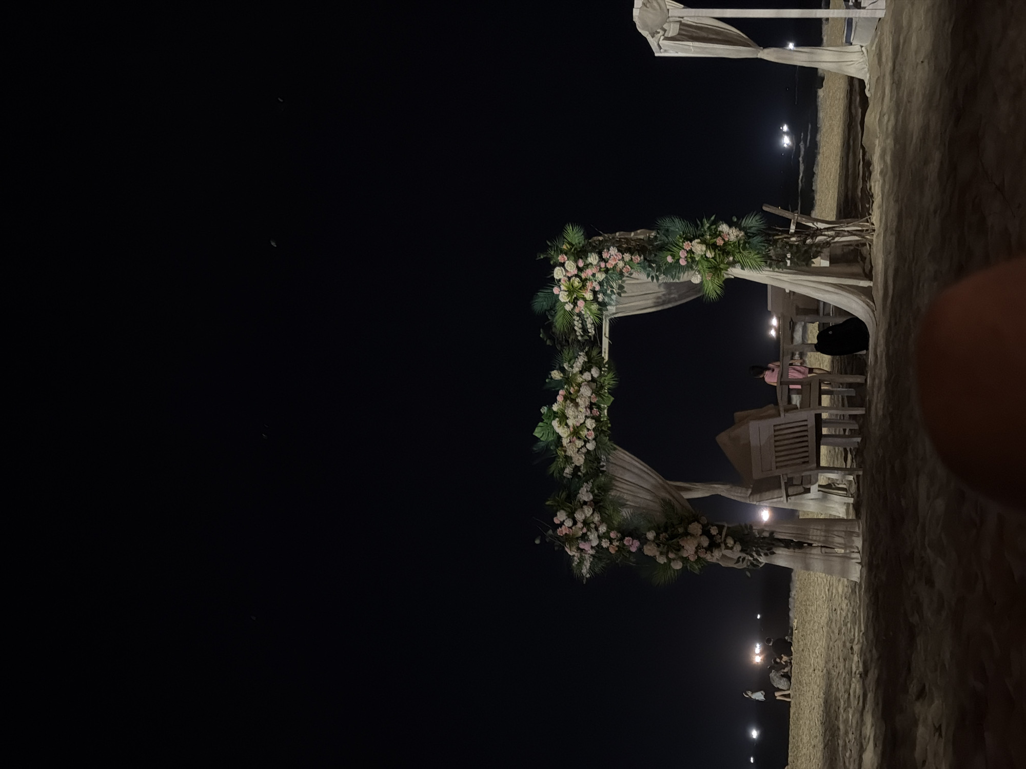 The image depicts a nighttime event set on a sandy beach in Đà Nẵng, Vietnam. The central focus is a beautifully decorated square archway, lavishly adorned with clusters of pink, white, and light green flowers, intermingled with vibrant green foliage, possibly palm fronds. The wooden structure of the archway is gracefully draped with light-colored, flowing fabric.
To the right of the archway, several wooden chairs and tables are visible. One person, wearing a light-colored top, is seated at a table, while another individual in dark clothing is seated further back. In the distance, several other people appear as dark silhouettes against the scattered lights illuminating the sandy ground. These numerous small, glowing lights create a warm and inviting ambiance across the beach.
The sky is completely dark, indicating a clear night, with no prominent stars or moon visible. The combination of the floral archway, arranged seating, and romantic lighting strongly suggests a special occasion, such as a beach wedding ceremony, a reception, or an elegant dinner event. The setting conveys a serene and celebratory atmosphere. No visible text is present in the media file.