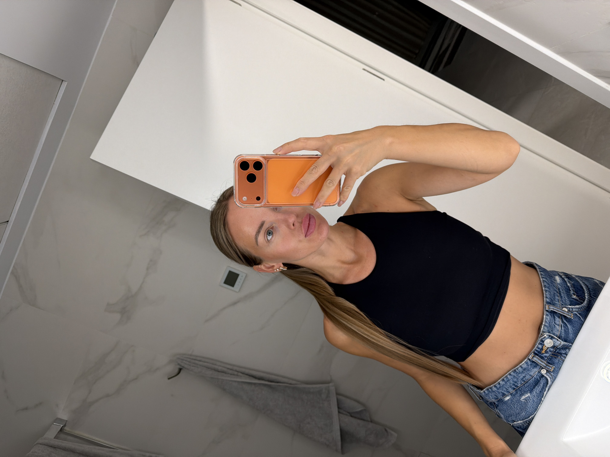 A woman with blonde hair tied in a ponytail takes a selfie in a bathroom. She is wearing a black crop top and denim jeans. She is holding an orange phone in her right hand. The bathroom has marble walls and a white sink. A grey towel hangs on a rack on the left side of the image. The image is shot from a slightly low angle.