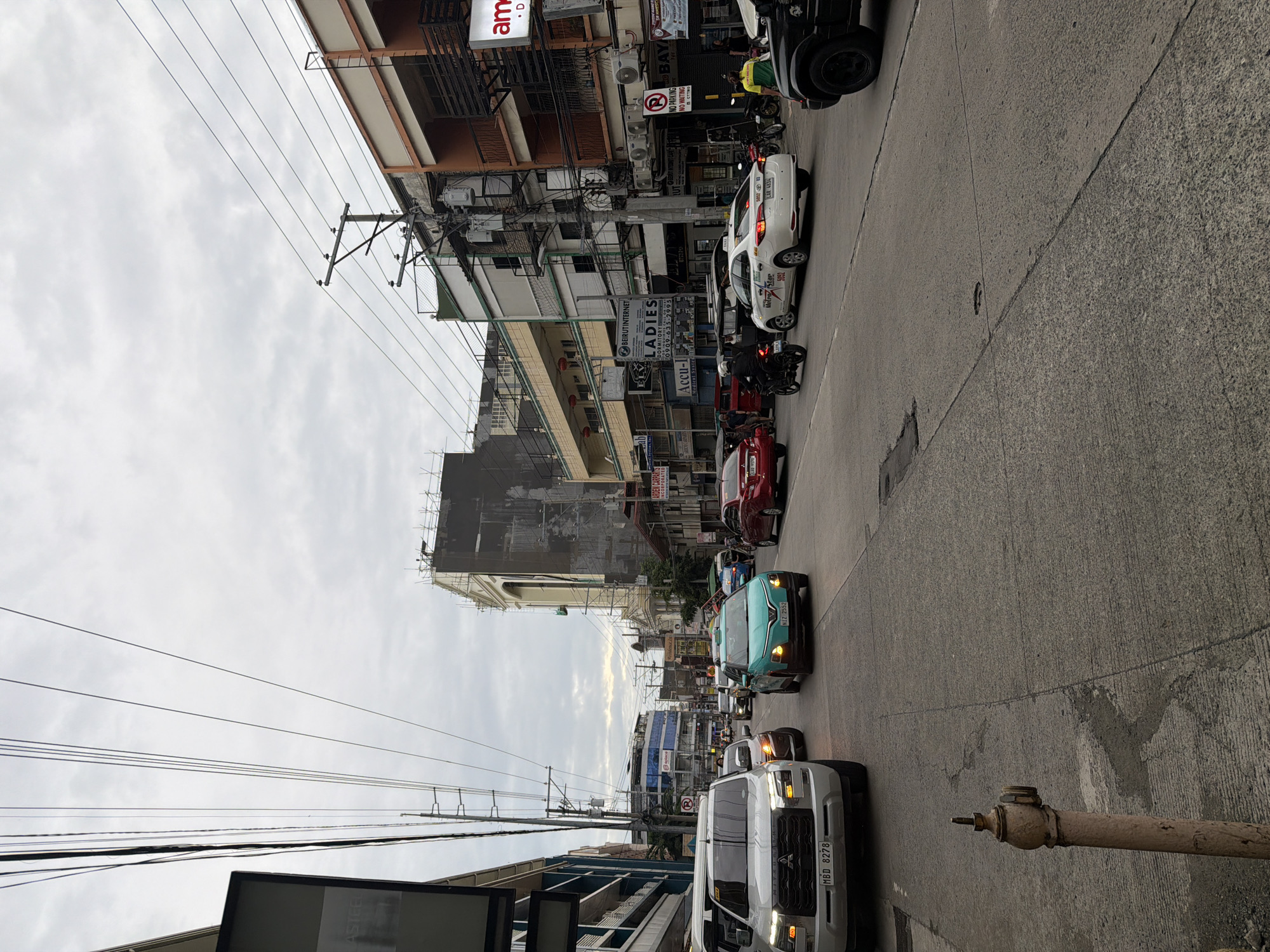 The image depicts a street scene in Davao City, Philippines, with multiple vehicles including cars, a truck, and motorcycles navigating the asphalt road. Power lines crisscross the cloudy sky above buildings lining the street. On the left side, a white truck with license plate "MBD 8278" is prominent. Further down the road, a turquoise car with license plate "NIZ 2552" is visible. Several buildings display various signs, including "BEIRUT INTERNET" and "LADIES DORMITORY." The environment appears to be urban, with signs of ongoing construction on one of the larger buildings in the background. The time of day seems to be late afternoon or early evening, given the lighting.
