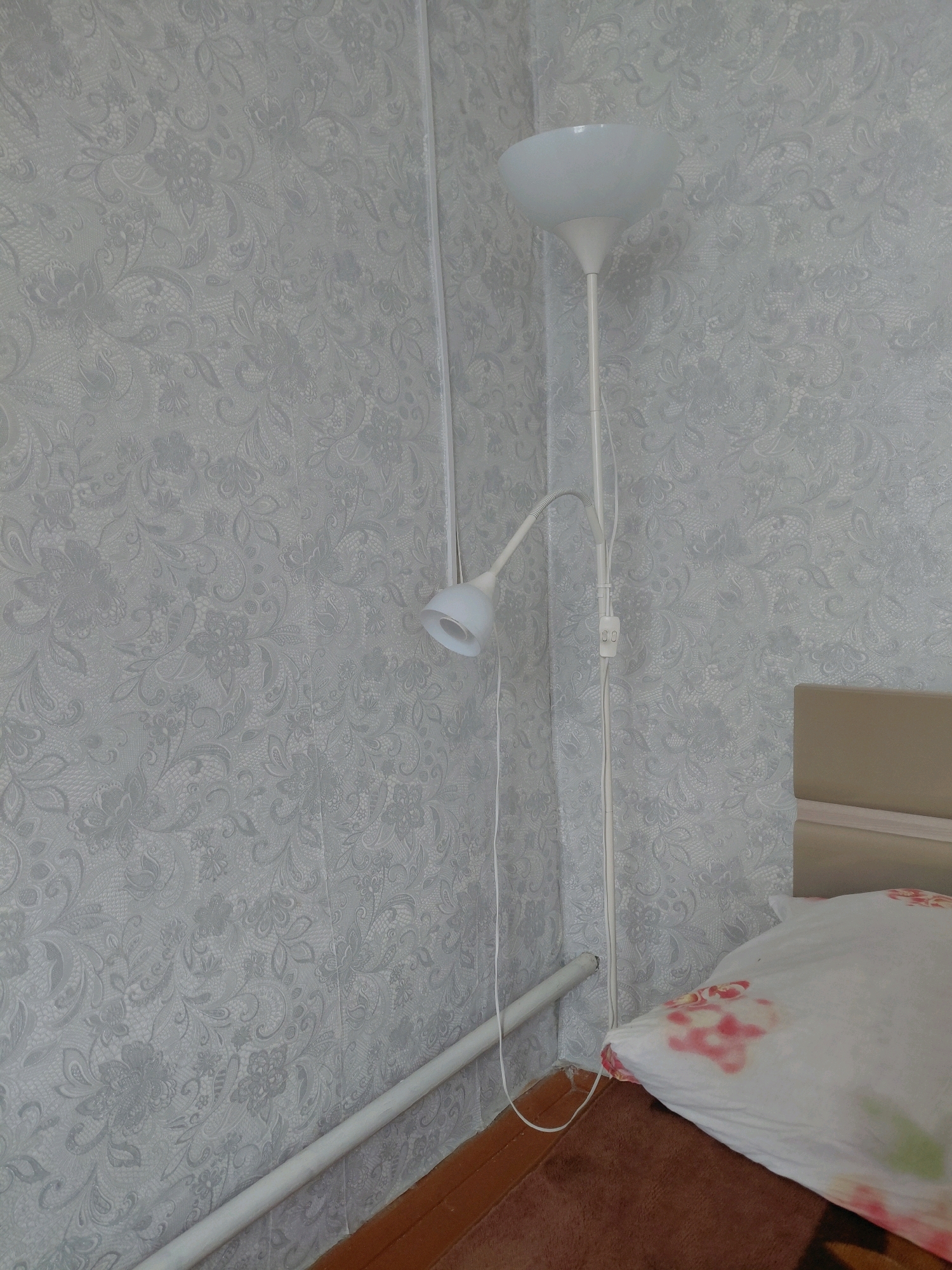A white floor lamp with a white shade stands in the corner of a room. The lamp has a flexible arm and a power cord with a switch. The wall is covered with light grey wallpaper with a floral pattern. A white pipe runs along the wall near the floor. To the right of the lamp, a pillow with a floral pattern lies on a bed. The flooring is brown. The scene appears to be an interior view of a room in Malinovka, Russia.