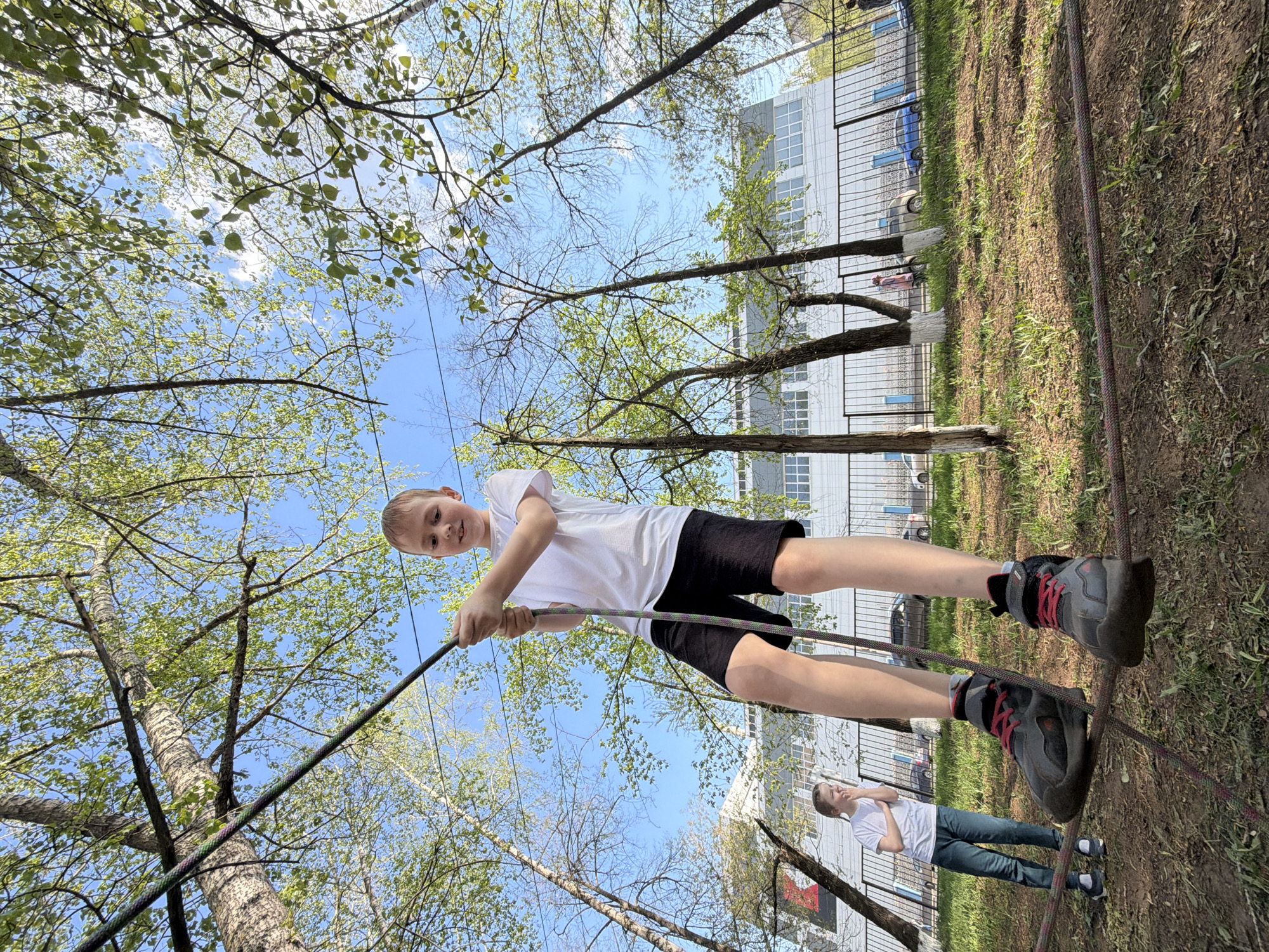 A low-angle shot shows a child (estimated age 9-12) on a rope climbing structure with another child (estimated age 12-15) watching. The first child is wearing a white t-shirt, black shorts, and hiking shoes. The second child is wearing a white t-shirt and blue jeans. The setting appears to be an outdoor recreation area in Izhevsk, Russia. A building in the background is a standard institutional-style building with a blue and white exterior. The image is taken on a sunny day with clear blue skies. The presence of spring foliage suggests the time of year.