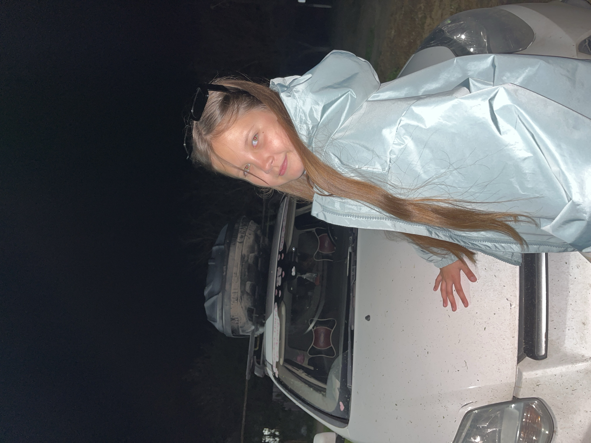 A young girl with long brown hair, wearing a light blue hooded jacket and sunglasses perched on her head, is leaning on the hood of a white vehicle. She is looking at the camera and her right hand is resting on the vehicle's hood. The vehicle appears to be dusty and possibly covered in debris. The background is dark, suggesting it is nighttime. The location is Novopokrovskaya, Russia.