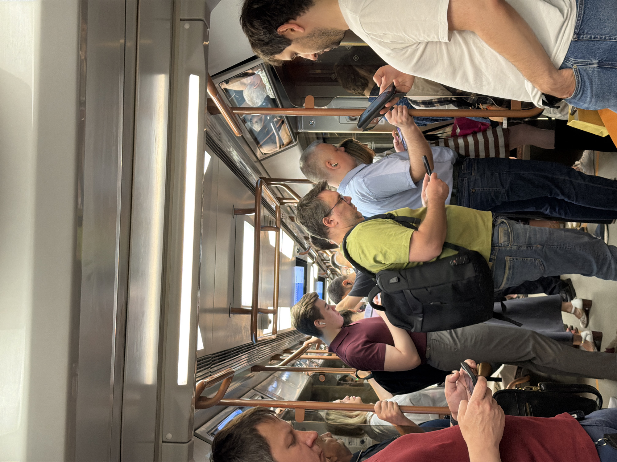 The image depicts a crowded train carriage, likely a metro, in Moscow, Russia. Passengers are standing, holding onto handrails, and looking at their mobile phones. The interior of the carriage is utilitarian with bright fluorescent lights and steel panels. The passengers are a diverse group in terms of age, dress, and ethnicity. Some are wearing casual clothing like t-shirts and jeans. A few are wearing backpacks or carrying bags.