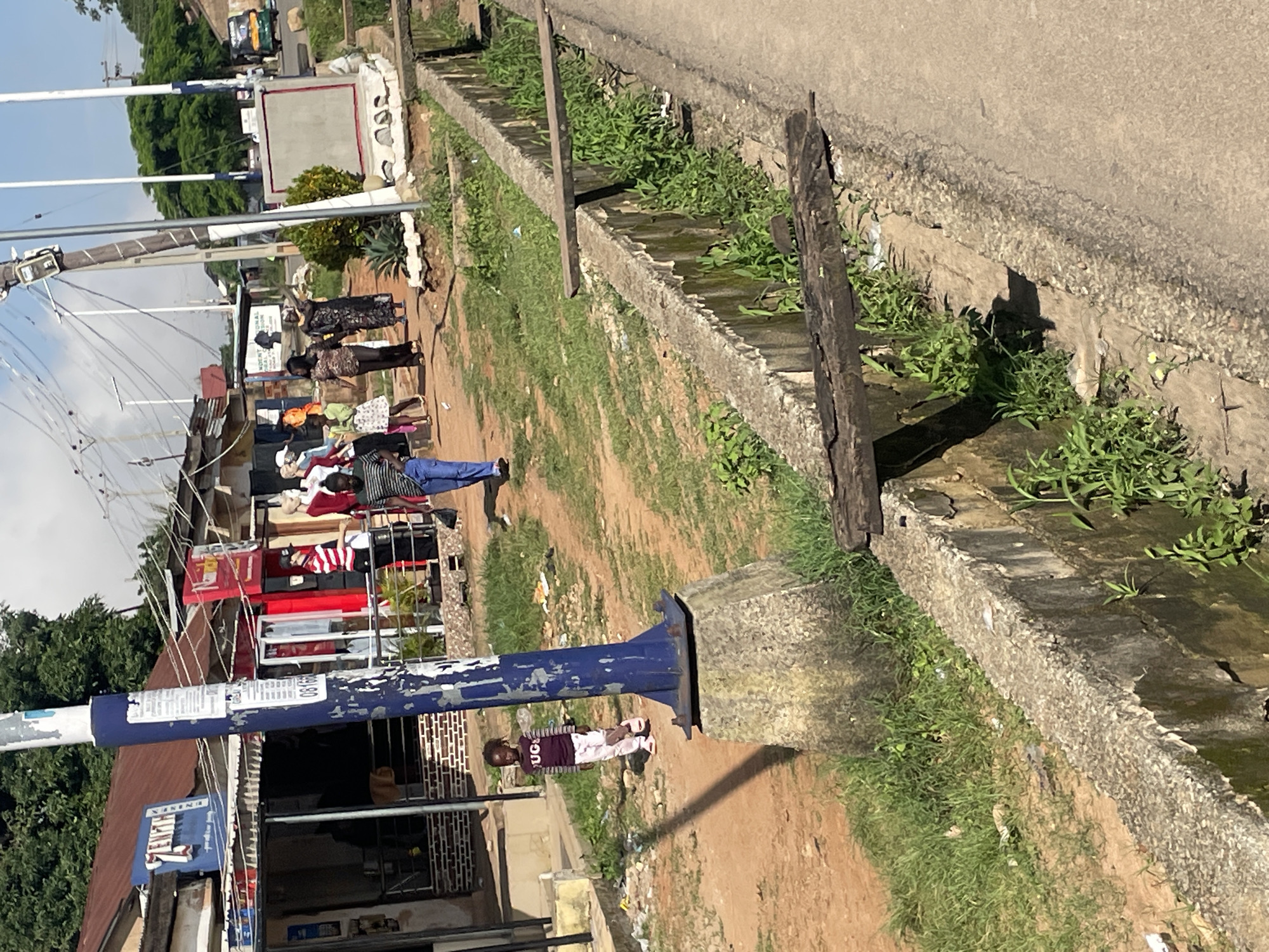 The image depicts a street scene in Jos, Nigeria. In the foreground, a concrete drainage channel runs along the side of a paved road. Wooden planks bridge the channel in several places. To the left of the drainage channel, a dirt path is visible, lined with grass and scattered debris. A blue metal pole, likely a utility pole, stands prominently on the dirt path.
Several individuals are present in the scene. A young child wearing a maroon long-sleeved shirt with the word "UGC" on it and white pants stands near the blue pole. Further down the dirt path, a group of people are walking. One individual, wearing a striped shirt and blue jeans, is walking towards the right side of the frame. Another person in the background is dressed in a black and patterned dress. Several mannequins are displayed outside shops on the left side of the image. These shops have corrugated metal roofs and open fronts. The background shows more buildings and vegetation, with power lines crisscrossing overhead. A yellow and green auto-rickshaw is visible on the road in the far distance. The overall environment suggests a commercial area in a developing urban setting.