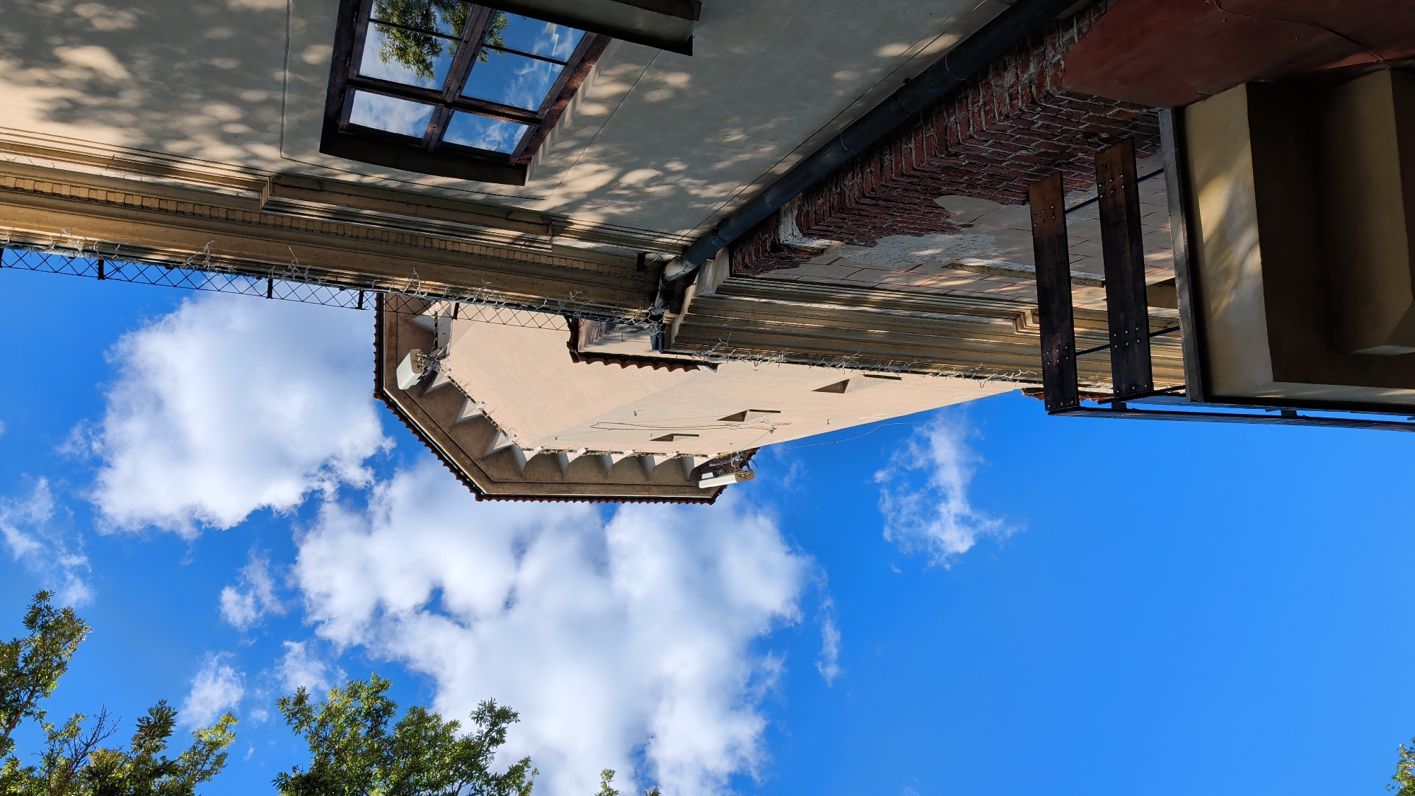 The image shows a low-angle view of an old building against a bright blue sky with scattered white clouds. The building's facade is light tan with decorative cornices and a tiled roof. A section of the building appears to have peeling plaster revealing red brick underneath. There is a dark downspout running vertically along the side of the building. A small, weathered wooden balcony with metal supports juts out from the building. A window with dark wooden frames and multiple panes is visible on the right side of the building, reflecting the sky and trees. A tree with green foliage is partially visible in the upper left corner. The image is oriented vertically, with the building occupying the right and lower portions of the frame. The lighting suggests it is daytime. The specific location in Pułtusk, Poland, is not determinable from the visual information provided in the image.
