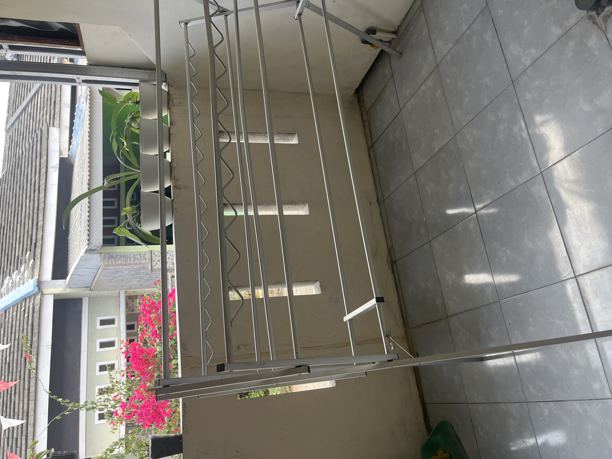 This image depicts an outdoor balcony area, likely in the city of Cirahayu, Indonesia, featuring a white metal clothes drying rack as the central subject. The rack is empty and positioned on a tiled floor. In the background, several potted plants are visible on a ledge, with a glimpse of houses and a roofline. The overall environment suggests a residential setting.