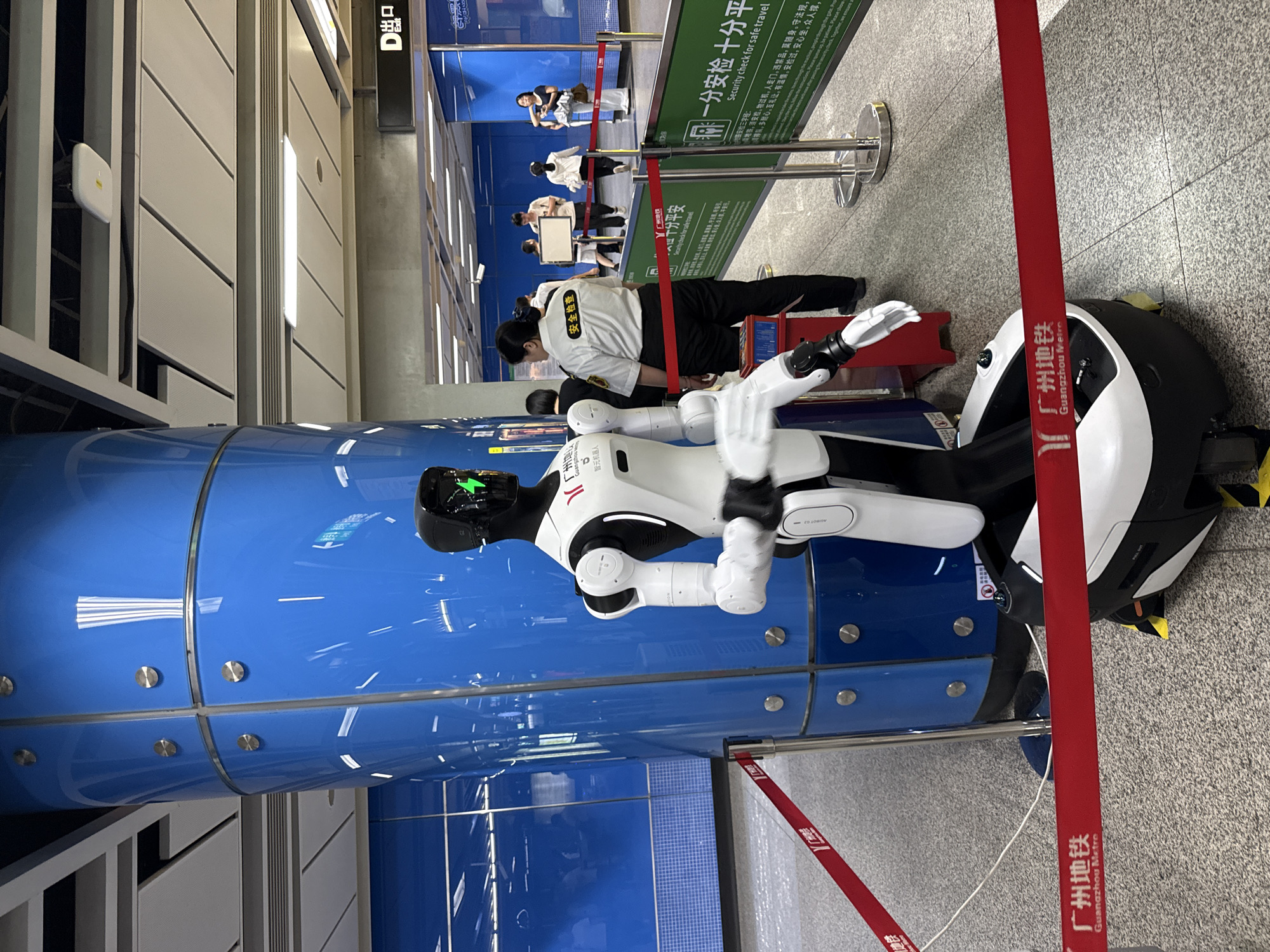 The image depicts a subway station in Haizhu District, China, featuring a prominent blue cylindrical pillar and a white and black humanoid robot. The robot stands to the right of the pillar, its face illuminated with a green light. Behind the robot, a security guard in a white uniform with "安全检查" (Security Check) written on the back observes the scene. Several other people are visible in the background, appearing to be passengers. Red velvet ropes are set up, indicating a designated area. Green signs with Chinese and English text, including "Security check for safe travel," are displayed. The environment appears to be a modern public transportation hub, with tiled flooring and a suspended ceiling.