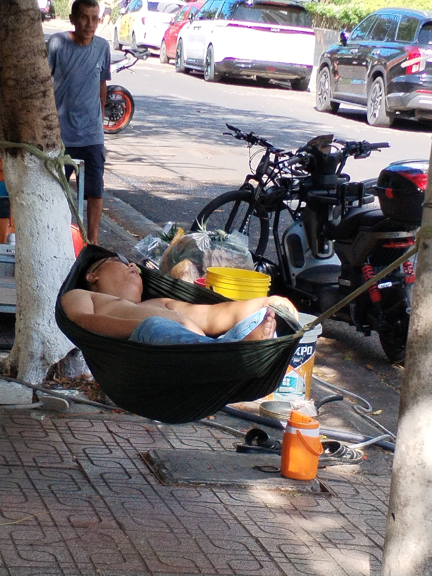 A man is sleeping shirtless in a green hammock, suspended between two trees on a sidewalk. He is wearing blue jeans and has glasses on his face. Another man stands nearby, looking at the person in the hammock. In the background, there is a street with parked cars, including a white KIA, a red car, and a black SUV. Next to the hammock, there is a bicycle, a scooter, a bucket of yellow paint, a can of paint, an orange water bottle, and a pair of sandals on the ground. The sidewalk is paved with textured brick-like tiles. The location is possibly in the city of Bac Nha Trang Ward, Vietnam.