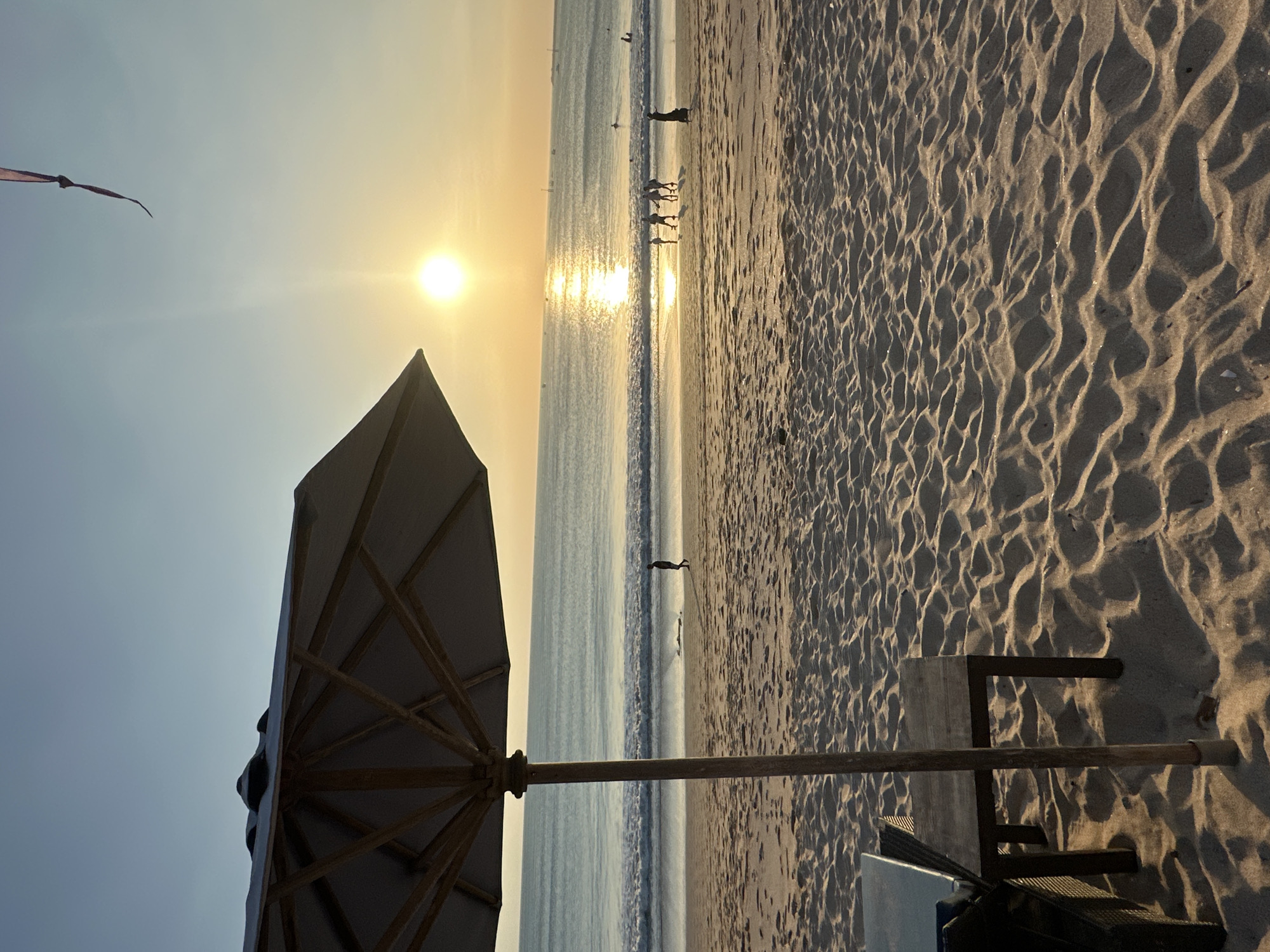 A wide-angle, eye-level shot captures a serene beach scene in Kuta, Indonesia, at sunset. In the foreground, a portion of a large, light-colored beach umbrella is visible on the left, its wooden frame and fabric canopy illuminated by the soft, warm light. To the right of the umbrella, a dark wooden table and chair sit on the sand, casting long shadows. The sand in the foreground is textured with ripples and undulations.
The middle ground features the gently rolling waves of the ocean meeting the sandy shore. Several people are scattered along the water's edge. A lone figure walks along the wet sand on the left. Further to the right, a group of four people are walking together near the water, and a solitary figure stands on the far right. In the distance, more figures are visible wading in the water, suggesting light activity.
The horizon is defined by the vast expanse of the ocean, stretching towards the setting sun. The sun, a bright, golden orb, is positioned slightly to the right of the center of the frame, casting a shimmering reflection on the water. The sky transitions from a pale blue near the top to a warm, golden hue closer to the horizon. A thin, purple ribbon-like object is visible in the upper right corner of the sky, possibly a kite string. Small, dark shapes, possibly boats, are visible on the horizon. The overall atmosphere is peaceful and tranquil, with the warm sunset colors dominating the scene.
