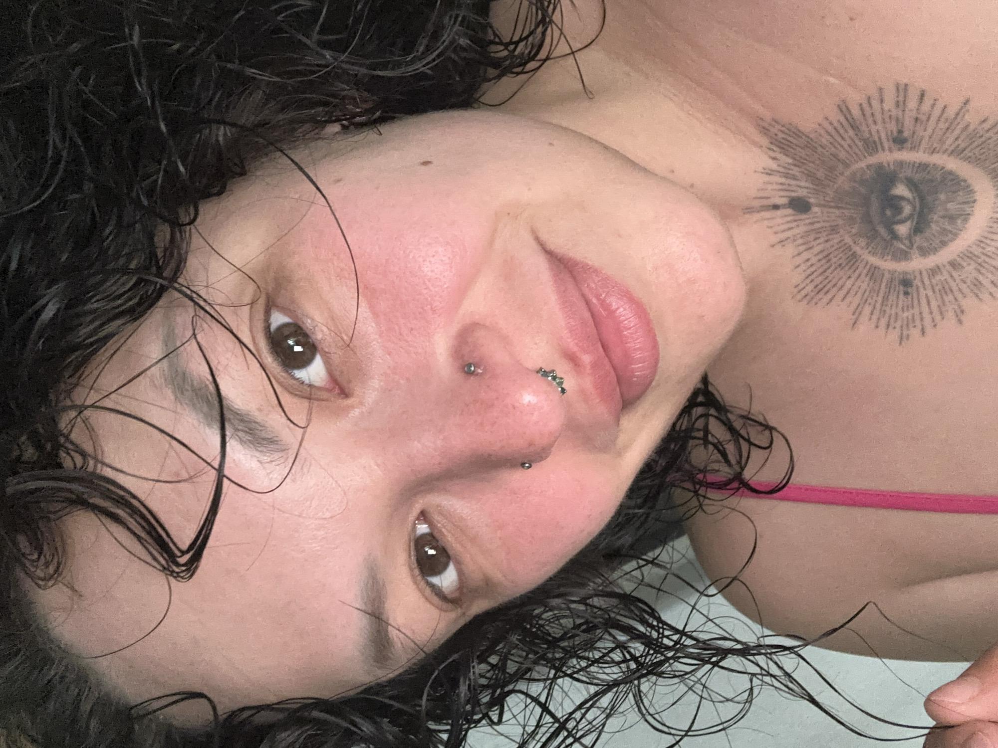 The image displays a close-up portrait of an individual, described as being located in the city of Chaikovsky, Russia. The subject has dark, wet, curly hair that partially obscures the forehead and shoulders. Their brown eyes are open and directed towards the viewer. The skin exhibits a light complexion with noticeable redness on the cheeks and nasal bridge. Two facial piercings are present: a silver-toned stud piercing on the upper central aspect of the nose and a cluster of small, dark-colored gems on the right nostril. The subject's lips are closed, forming a subtle smile. A prominent tattoo depicting a stylized eye surrounded by radiating lines and small dots is visible on the upper sternum/chest area. A single pink strap from a garment, possibly swimwear or an undergarment, is visible over the subject's right shoulder (viewer's left). The background is an undifferentiated, light-toned surface.