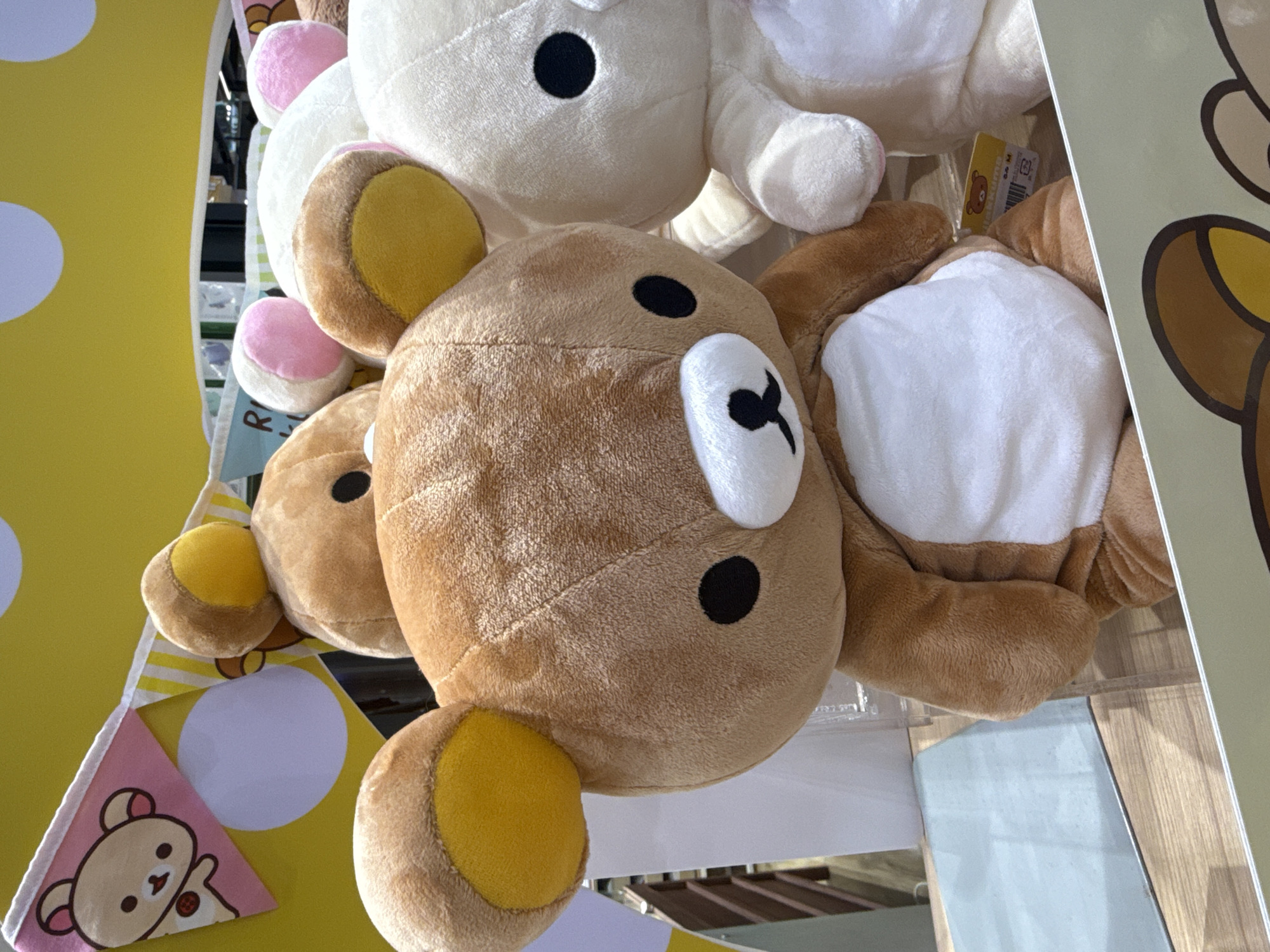 This is a close-up shot of several stuffed animals, possibly in a retail display. In the foreground, a large, brown teddy bear with yellow inner ears is the primary subject. It has black button eyes and a black stitched mouth and nose. Its belly is a patch of white fabric. Behind it, to the right, is a white stuffed animal with black button eyes and pink inner ears. Other stuffed animal heads are visible in the background, including one with pink ears. A yellow banner with white polka dots and a pink triangular flag featuring a cartoon bear face is visible at the top left of the image. The stuffed animals appear to be on a wooden surface, and there is some reflective material, possibly glass or plastic, in the background which shows indistinct shapes and lighting. The overall impression is of a brightly lit, cheerful display.
While the image depicts a scene that could be found in a store anywhere, the prompt requests specific location information for Bangkok, Thailand. Without additional information, it is impossible to definitively place this image in Bangkok, Thailand. However, if this image was taken in Bangkok, it could be in a toy store or a department store in a shopping mall such as Siam Paragon, CentralWorld, or ICONSIAM.