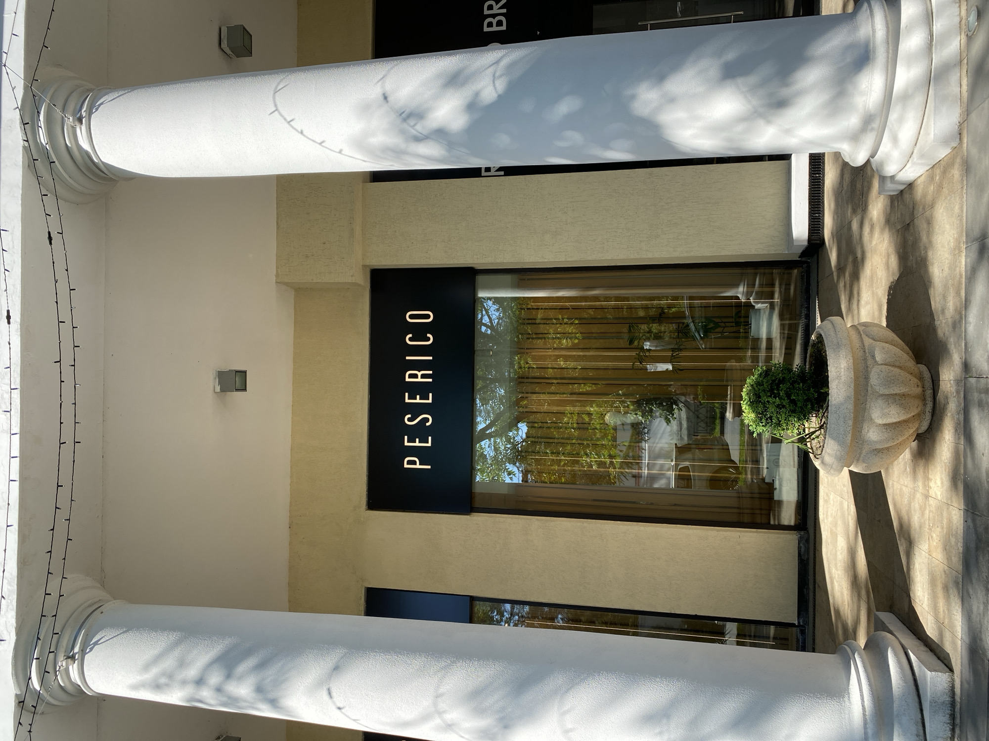 The image displays the entrance of a building in Sochi, Russia, characterized by classical architecture and modern storefront design. Two substantial white columns, with fluted bases and a slightly textured surface, frame the view in the foreground.
Bright sunlight strongly illuminates the scene, creating sharp shadows of leafy tree branches on the columns and the light-colored tiled pavement. The building facade features creamy-tan, textured walls. Centrally positioned is a store entrance with a large, rectangular black sign displaying the brand name "PESERICO" in white, vertical capital letters. Below this sign, a wide glass window reflects the surrounding environment, including green trees and what appears to be a clear sky. Inside the store, warm-toned curtains are visible through the reflection.
To the right of the window, a decorative, ornate stone planter holds a small, well-manicured green bush. On the left column, a string of small, dark-colored fairy lights hangs vertically, adding a subtle decorative element. Higher up on the building, partially obscured by the right column and the image crop, additional white text on a black background reads "R BR," likely part of a larger sign.
The scene depicts a clear, sunny day. No people or specific activities are present, focusing attention on the architectural details and the retail establishment. The overall impression is one of a clean, well-maintained, and possibly upscale commercial space.