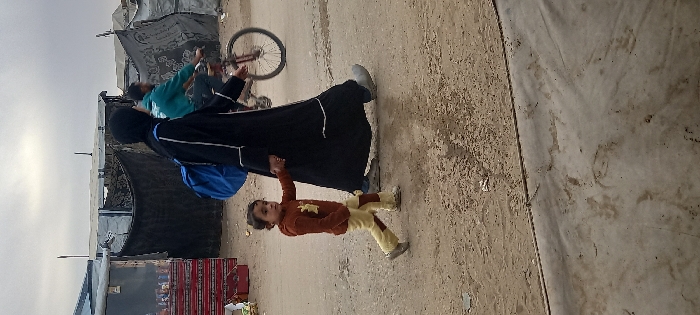 A woman in a black robe walks on a dirt path, holding the hand of a young child in a brown sweater and light yellow pants. Behind them, a person on a bicycle is visible amidst makeshift shelters and tents. A blue backpack is visible on the woman's back. The surroundings appear to be a refugee or displaced persons camp.