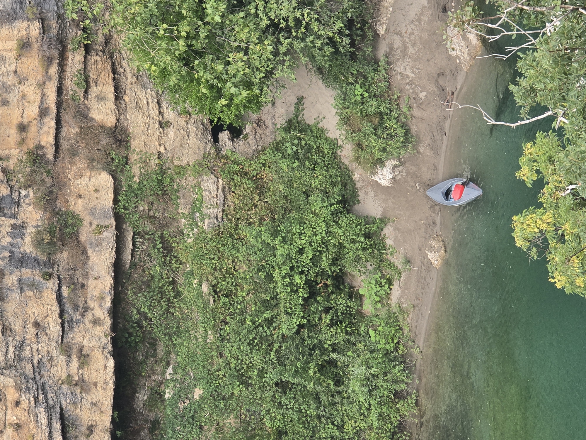 A high-angle view shows a canoe with a red object inside resting on the shore of a clear, turquoise body of water. The water's edge is lined with a sandy bank covered in sparse vegetation. Behind the bank, a dense wall of green foliage rises, obscuring the base of a rugged, layered rock formation that dominates the upper portion of the frame. The rock face is a mix of tan and grey, with visible striations and patches of dry vegetation. A dark opening, possibly a cave entrance, is visible within the rock face, partially concealed by the surrounding greenery. The overall scene suggests a natural, undeveloped environment.