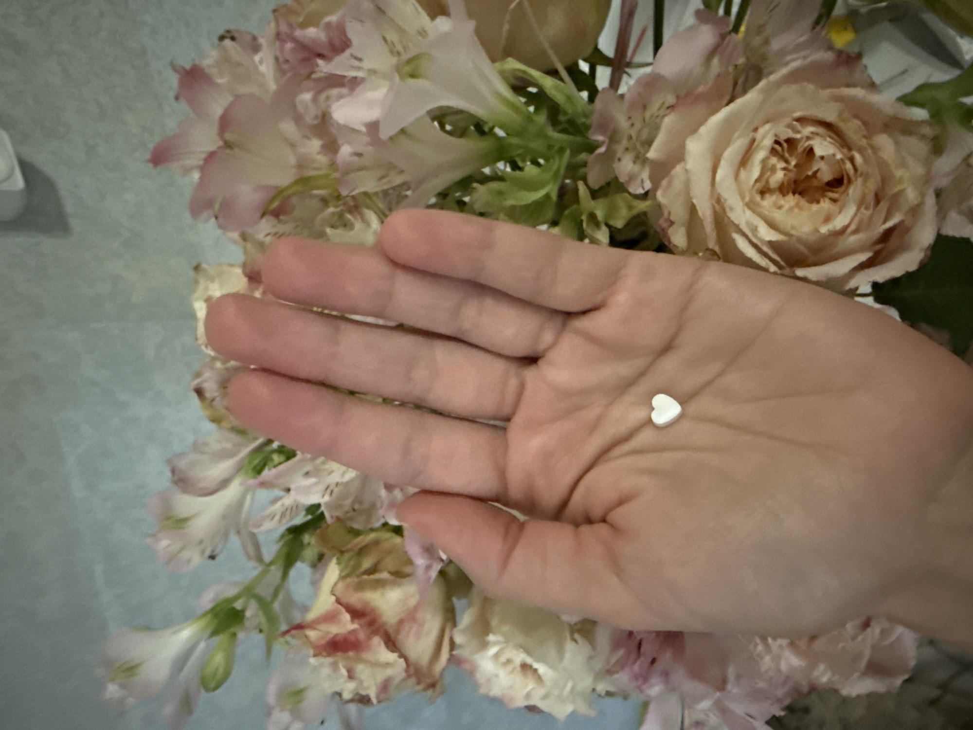 A close-up, overhead shot shows a person's open palm against a backdrop of a bouquet of pink and white flowers. A tiny, white, heart-shaped object rests in the center of the palm. The background is out of focus, highlighting the hand and the small heart. The image appears to have been taken indoors, with the lighting suggesting it is daytime. There are no visible landmarks or contextual clues to determine the location within Saint Petersburg, Russia.