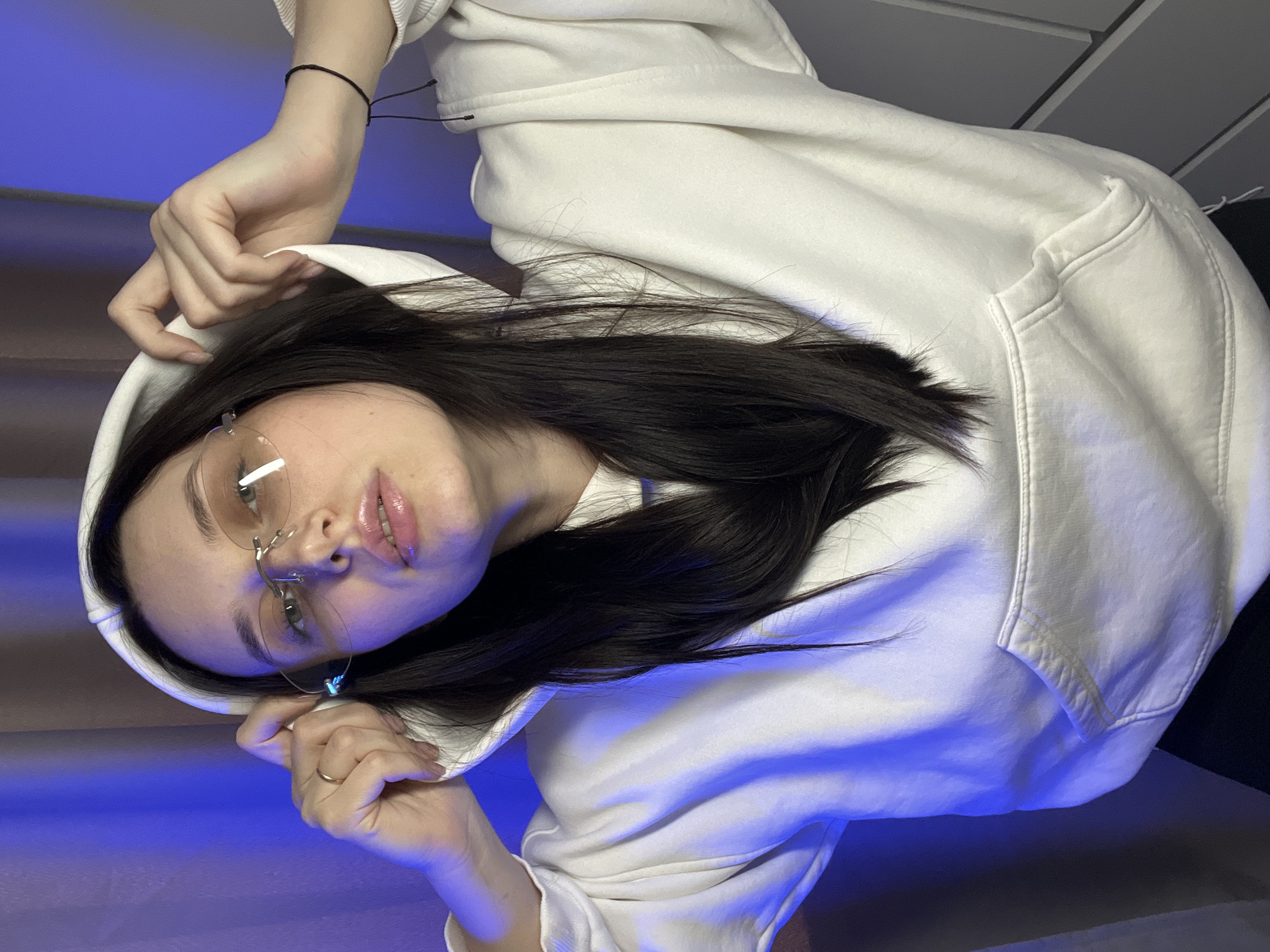 An indoor, close-up portrait shot features one individual, identified as female, positioned centrally. The subject has dark, straight hair, wearing a white hooded sweatshirt with a prominent front kangaroo pocket. She is also wearing rimless eyeglasses with light brown or tinted lenses. A gold-colored ring is visible on the fourth finger of her left hand, which is raised, along with her right hand, to hold or adjust the hood of the sweatshirt. The nails on both hands appear to have light-colored polish. A thin black cord bracelet is worn on her right wrist. Her mouth is slightly open, revealing what appears to be a lip piercing on the lower lip. The background on the left consists of a light brown or tan curtain/fabric, partially obscured and illuminated by a strong blue light source originating from the left, casting a distinct blue hue onto the subject's left side and the adjacent background. The background on the right shows a light gray wall or furniture with horizontal paneling or drawer lines. The specific location within the city of Tyumen, Russia, cannot be determined from the visual information presented in this image.