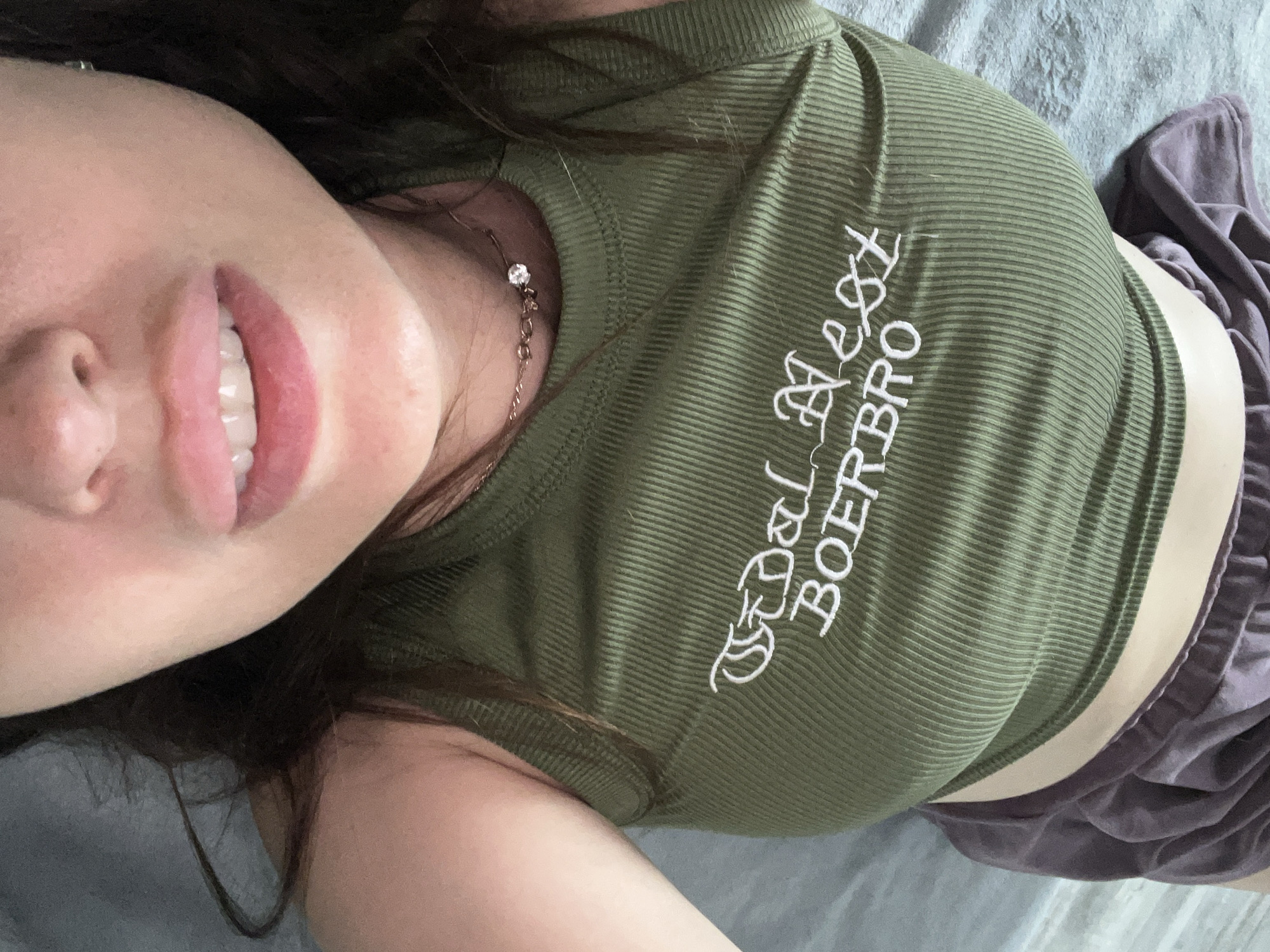 An overhead shot captures a person lying on a bed, facing upwards. They are wearing a green, ribbed tank top with white embroidery that reads "WEST BOERBRO" and a pair of purple shorts. A delicate necklace with a small pendant rests on their collarbone. The person's mouth is slightly open, revealing their teeth, and their dark hair is spread around their face. The background consists of a blue, textured bedsheet.