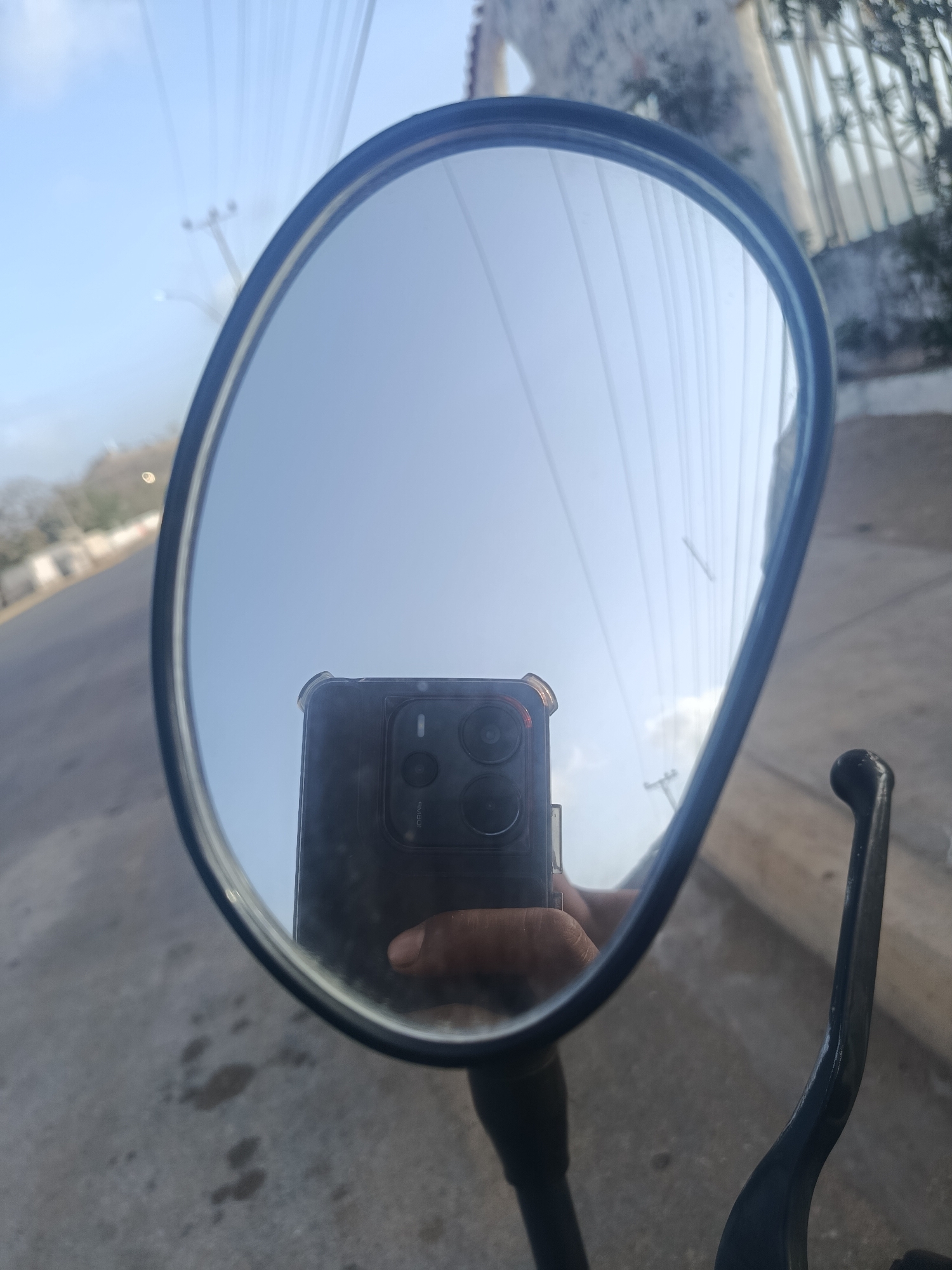 The image is a reflection in a motorcycle mirror of a hand holding a smartphone, apparently taking a selfie. The background in the mirror includes power lines, the sky, a building, and trees. Outside the mirror, parts of the motorcycle, including a handlebar and a brake lever, are visible. The location, based on the prompt, is Juan Griego, Venezuela, but specific details confirming the location cannot be extracted directly from the visual data.