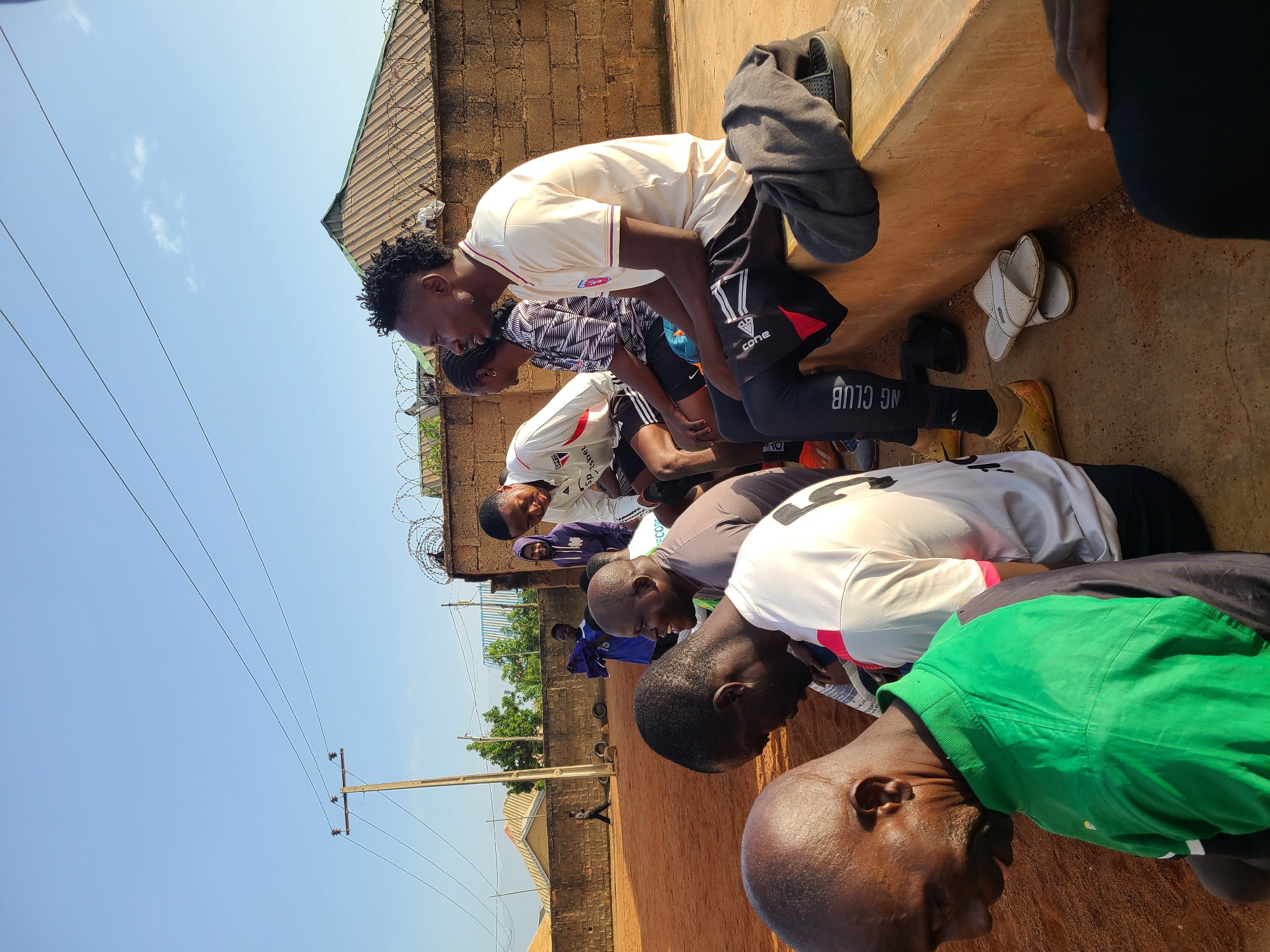 The image shows a group of men and one woman sitting on a concrete ledge outdoors. The setting appears to be a sports field, with red dirt visible in the foreground. In the background, there's a brick wall topped with barbed wire, a building with a corrugated roof, and utility poles with wires. The sky is blue and clear.
The individuals are casually dressed, some in athletic wear. Some men are looking at each other or in different directions, while others appear to be resting. One man in the foreground is wearing a green and black jacket, and a man next to him is wearing a white and grey athletic shirt. A woman with braided hair is seated to the right of a man in a light-colored polo shirt. Some pairs of sandals are on the ground near the ledge. The overall impression is a pause during an outdoor activity or gathering.