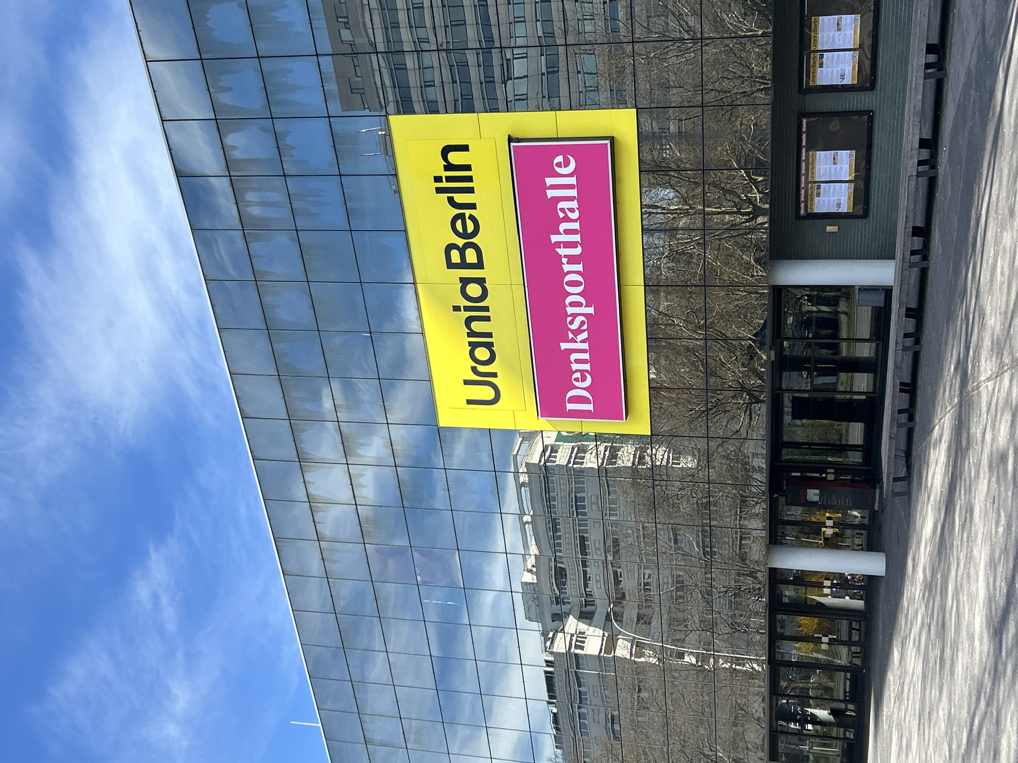 The image shows the Urania Berlin building, a cultural center in Berlin, Germany. The building has a modern glass facade reflecting the sky and nearby buildings. A sign on the building reads "Urania Berlin" in yellow and "Denksporthalle" in pink. The entrance has glass doors and columns. Posters can be seen on the side. The area appears to be a public space, with a sidewalk.