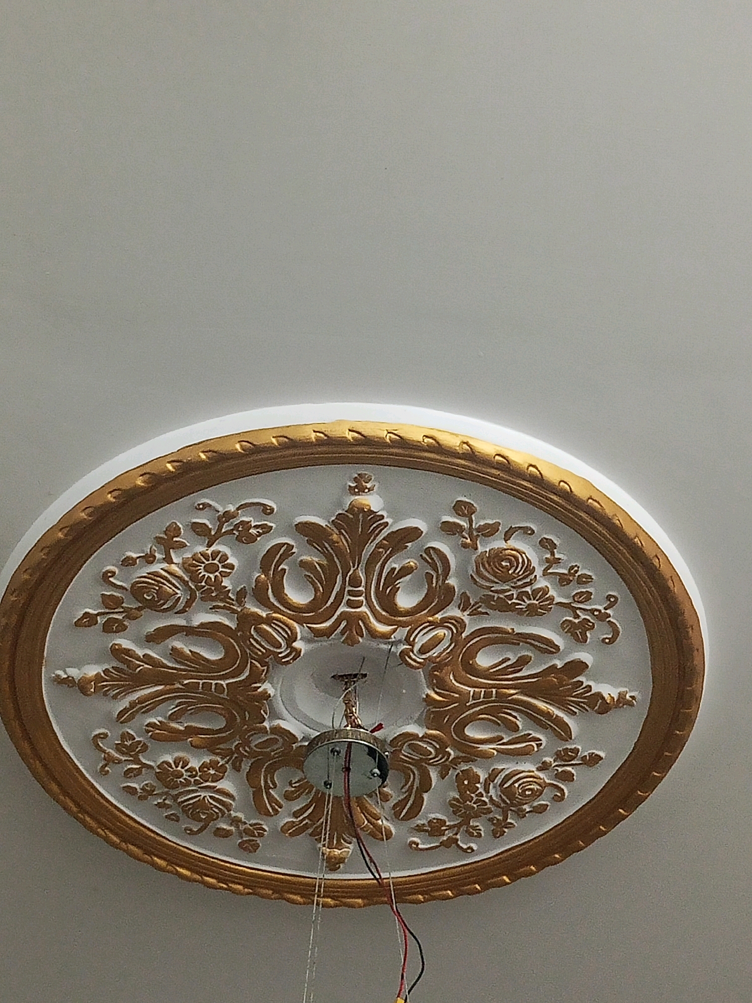 The image shows a white and gold ornate ceiling medallion with floral and scrollwork designs. A light fixture with wires is attached to the center of the medallion. The medallion is mounted on a white ceiling. The photo was taken in Bhedian - Nai Basti, Pakistan.
