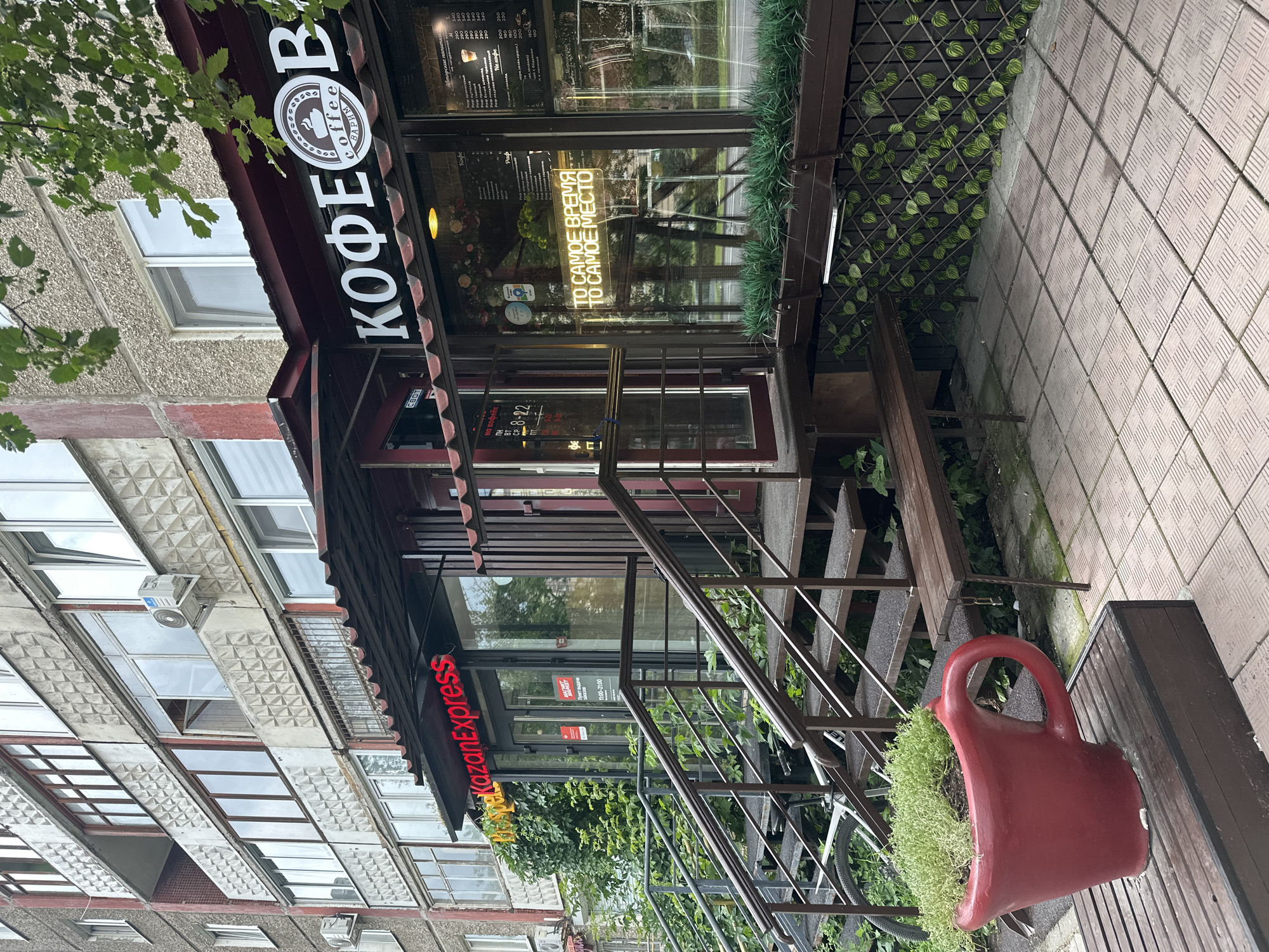 This is an exterior, medium-shot, eye-level, daytime photograph of a storefront in the city of Perm, Russia. The storefront, likely a cafe or coffee shop, is situated on the ground floor of a multi-story apartment building with a grey, textured facade and numerous windows. The building appears to be older, with some visible signs of wear and tear.
The storefront features a dark red awning extending over the entrance. To the left, a neon red sign reads "KazanExpress." To the right, a large white sign above the window displays the word "КОФЕОВ" in Cyrillic, with a circular logo featuring the word "coffee" and "варим" (meaning "we brew") beneath it. Below this, a neon sign in yellow letters reads "ТО САМОЕ ВРЕМЯ TO САМОЕ МЕСТО" (which translates to "The Right Time, The Right Place").
A set of metal stairs with handrails leads up to the entrance. The stairs are flanked by overgrown green plants. In the foreground, to the left, is a large, bright red planter shaped like a coffee cup, filled with green plants. A wooden bench is visible to the right of the planter. The ground in front of the storefront is paved with square tiles. The overall scene suggests a casual, urban environment.