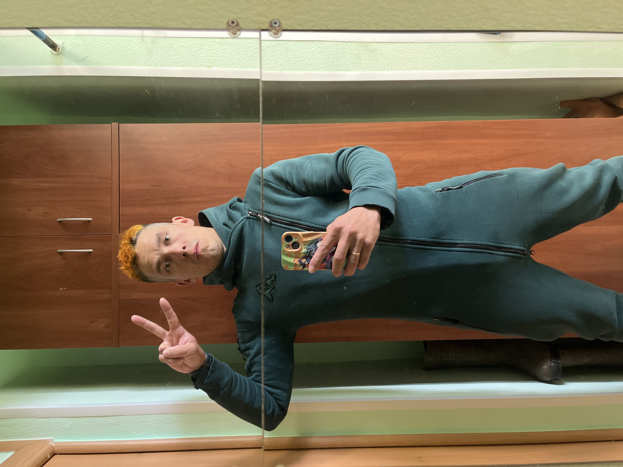 A man with orange hair is taking a selfie in a mirror. He is wearing a dark green jumpsuit. He is holding a cell phone in his right hand and making a peace sign with his left hand. The background includes a wooden cabinet, a green wall, and what appears to be a shelf with wooden items. The location is Chaikovsky, Russia.