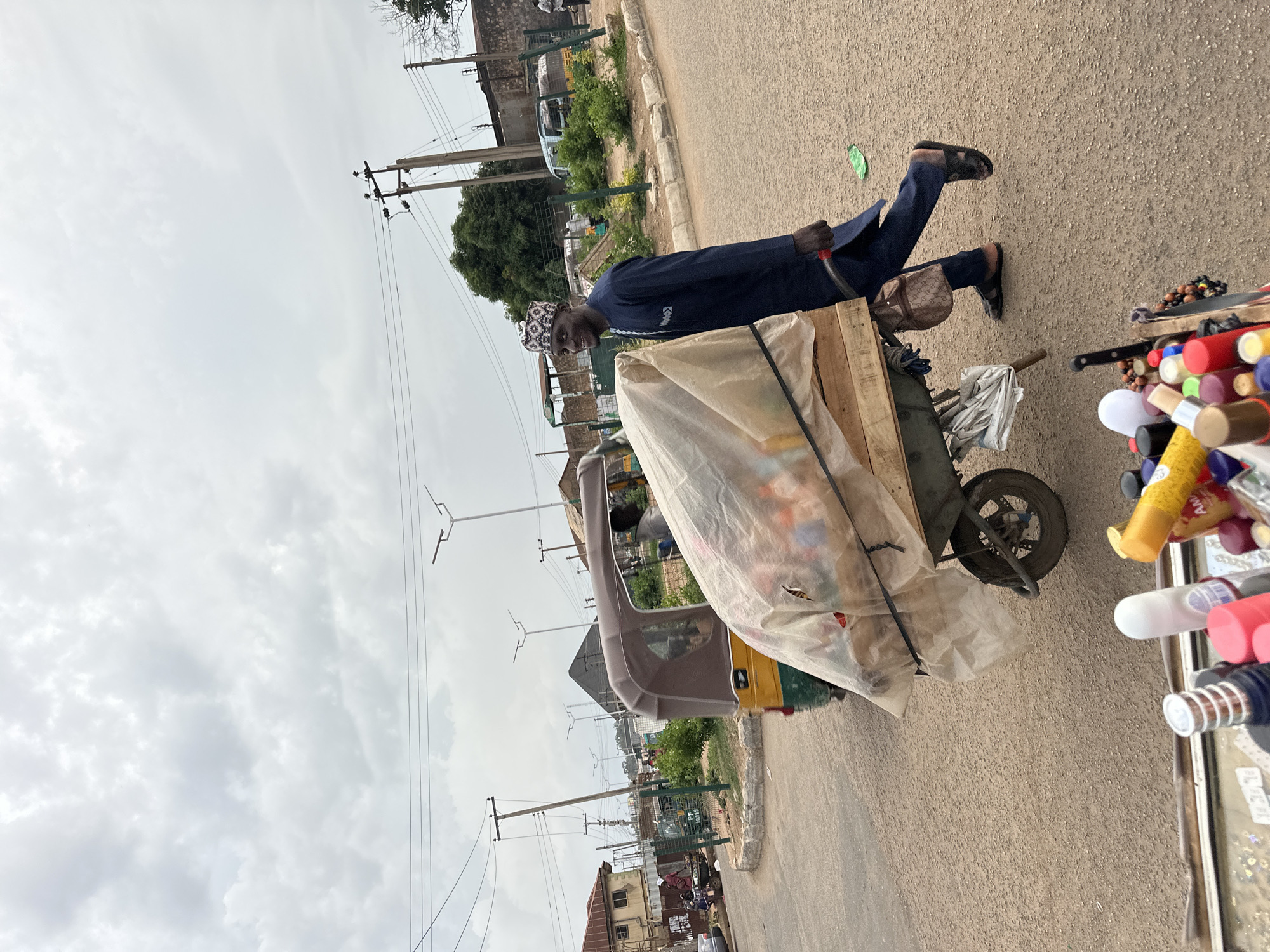 The image depicts a street scene in Bukuru, Nigeria. A man wearing a dark blue traditional garment and a patterned cap is walking beside a wheelbarrow laden with goods covered by a translucent sheet. The wheelbarrow is made of wood and metal and has a single wheel. Behind the man and the wheelbarrow, a yellow and green auto-rickshaw is visible. Further in the background, there are buildings, trees, utility poles with wires, and other vehicles, including another auto-rickshaw and a white minivan. The ground is paved with asphalt, and the sky is overcast with grey clouds. In the foreground, the tops of various bottles and containers are visible, suggesting a market stall or personal items. The man appears to be smiling and looking towards the viewer. The overall environment suggests a busy urban or semi-urban setting.