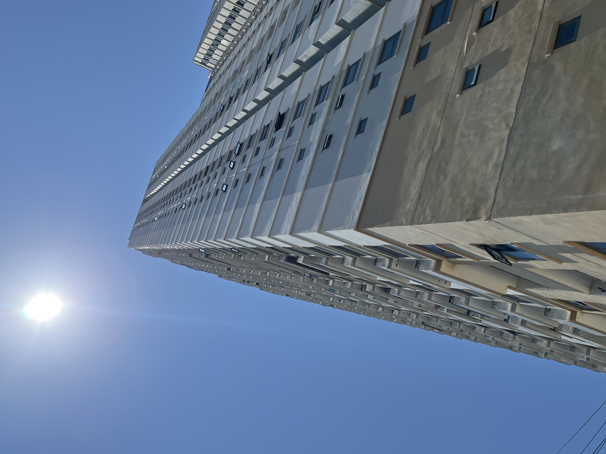 The image shows a tall, multi-story building from a low angle. The building has a modern design with many rectangular windows. Some windows are open. The building is likely a residential or commercial structure. The sky is clear and blue. The sun is visible in the lower left corner of the frame. Location in the city of Đà Nẵng, Vietnam is not evidenced.