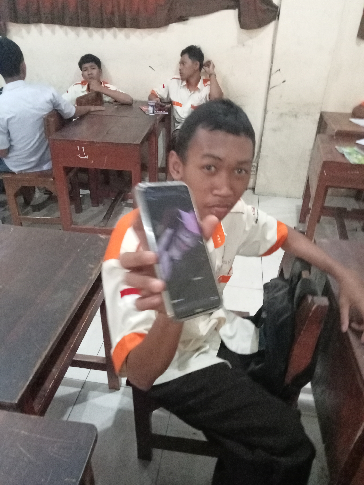 The image depicts a classroom setting at a school in Secang, Indonesia. In the foreground, a young male student is holding up a smartphone, appearing to take a photo or video of himself. He is wearing a light-colored shirt with orange trim and dark pants. He is seated at a wooden desk, and a backpack is visible behind him. In the background, other students are seated at desks, some of whom are also wearing similar school uniforms. The room has simple wooden furniture and a light-colored wall. The overall impression is of a casual moment captured during a school day.