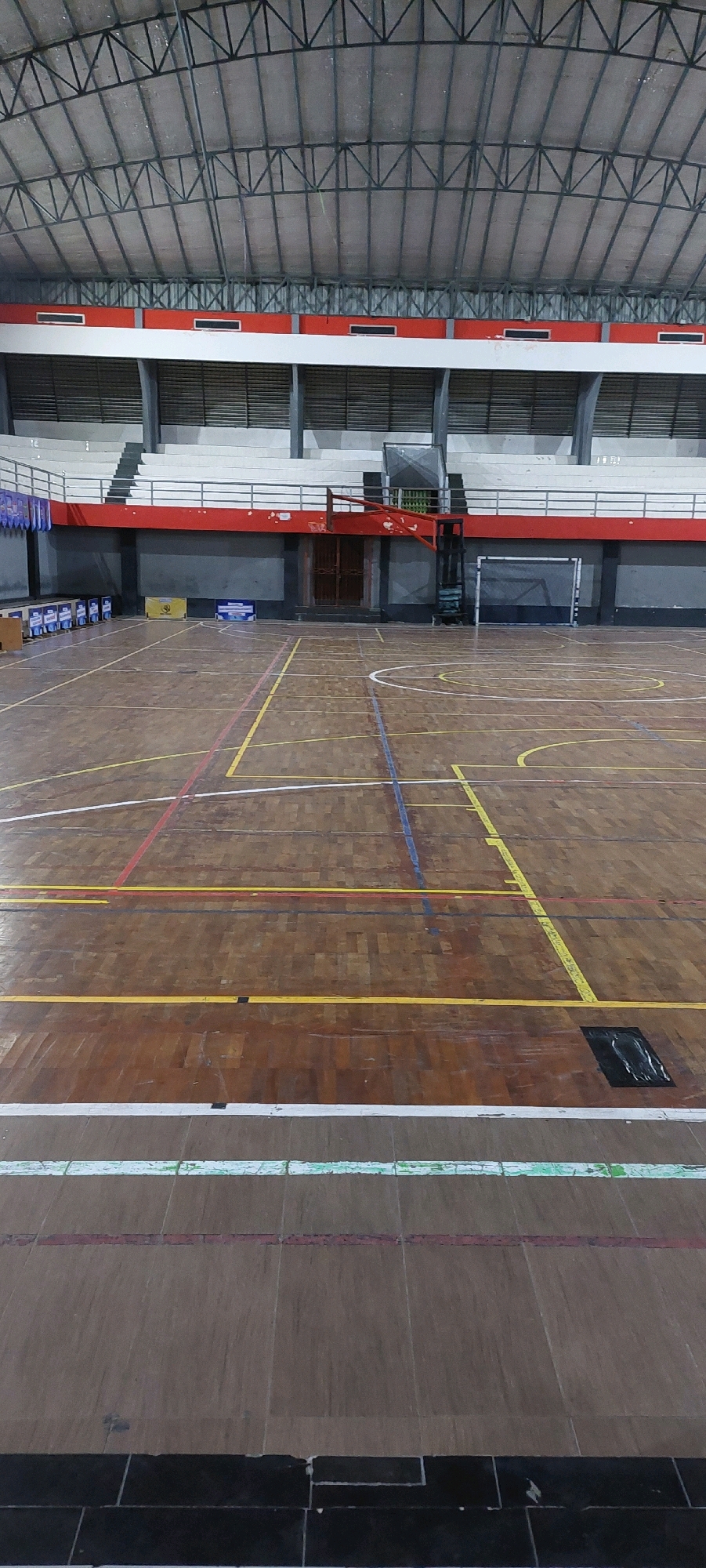An interior view of an empty sports gymnasium is captured. The gymnasium features a wooden floor with various colored lines marking the boundaries of different sports courts. In the background, tiered seating is visible, with a red railing and white benches. The ceiling is a high, arched structure supported by metal trusses. On the right side of the court, a basketball hoop and backboard are mounted, and further back, a soccer goal net is visible. The overall lighting suggests it is daytime, likely with artificial lighting from the gymnasium's fixtures. The scene appears to be in an unspecified location in an unknown city.