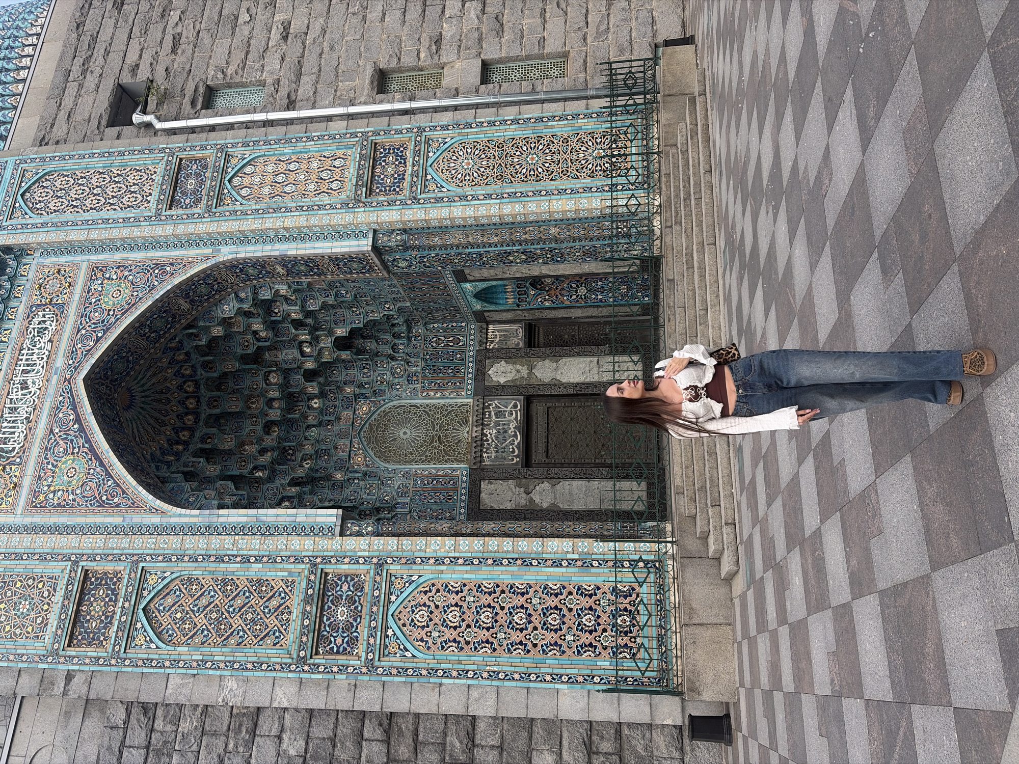 A young woman stands in front of the Saint Petersburg Mosque. The mosque is made of granite with intricate blue and gold tilework adorning the entrance. The woman is wearing a white cropped sweater, a brown camisole, and blue jeans. She has a leopard print purse slung over her shoulder. The ground in front of the mosque is paved with a checkered pattern of gray and darker gray tiles. The sky is overcast.