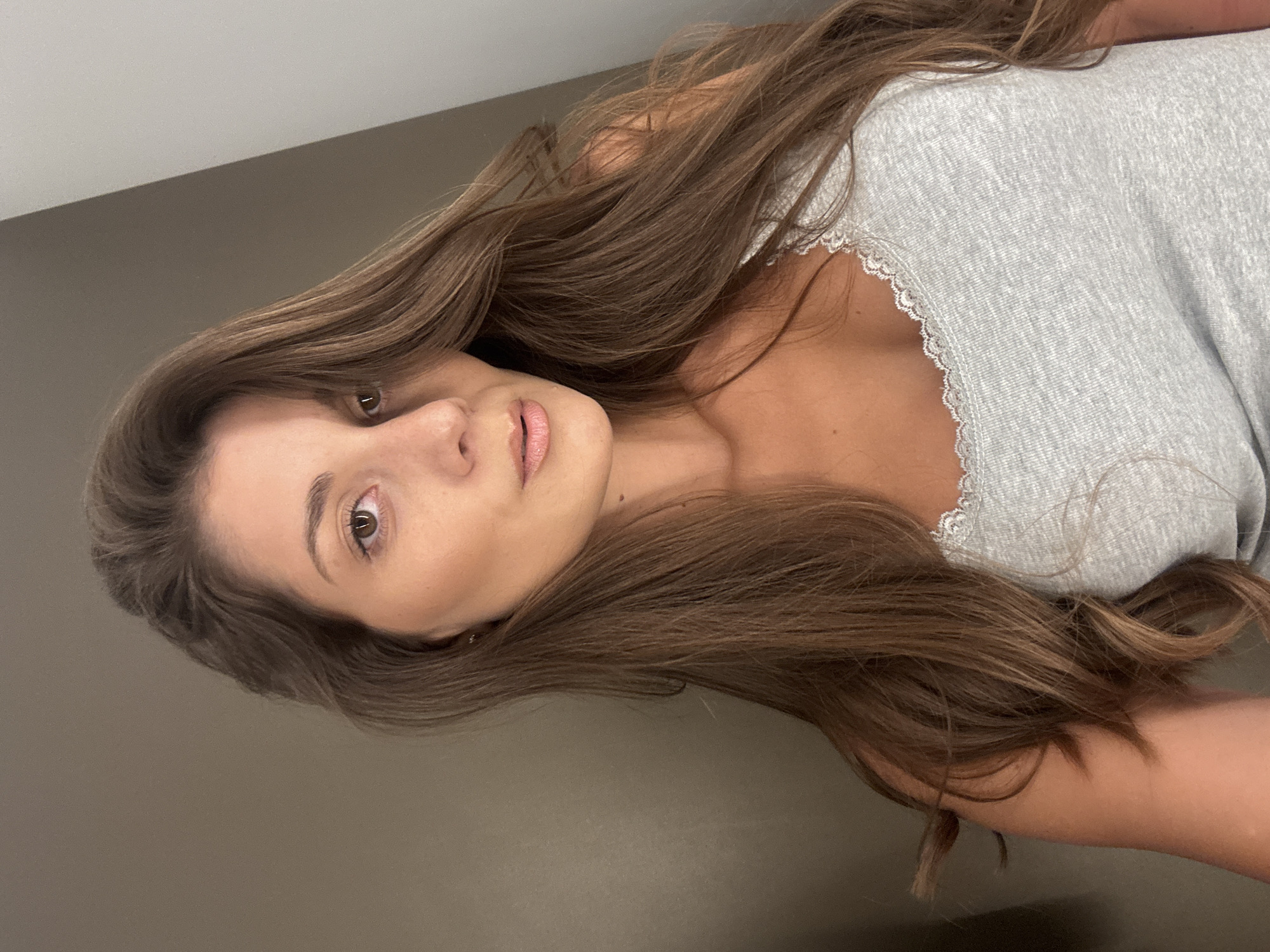 A portrait of a woman with long brown hair and fair skin is captured. She is wearing a grey top with lace detailing around the neckline. The background appears to be a plain, neutral-toned wall. There is no evidence in the image to determine that the location is in Burgas, Bulgaria.